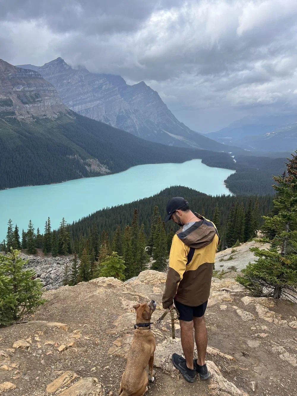 Trey Foster standing on a rocky overlook with a brown dog, above a turquoise mountain lake in the Rockies
