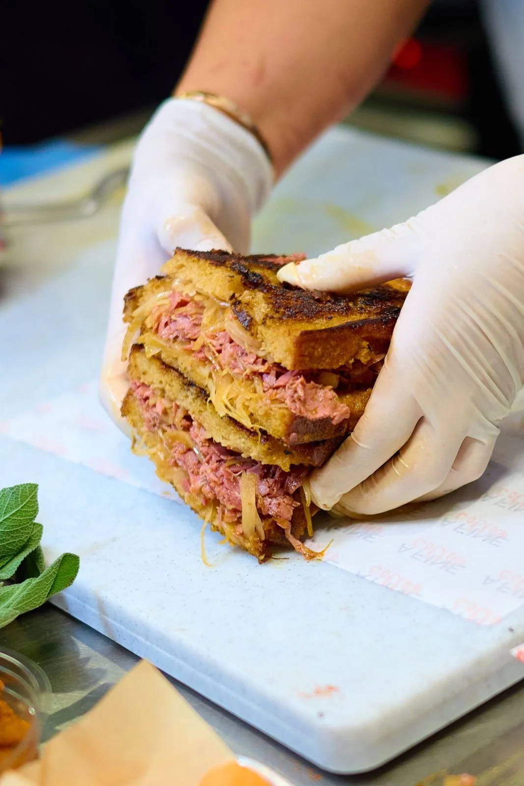 Personnes portant des gants préparant un sandwich au pastrami avec du fromage fondu.