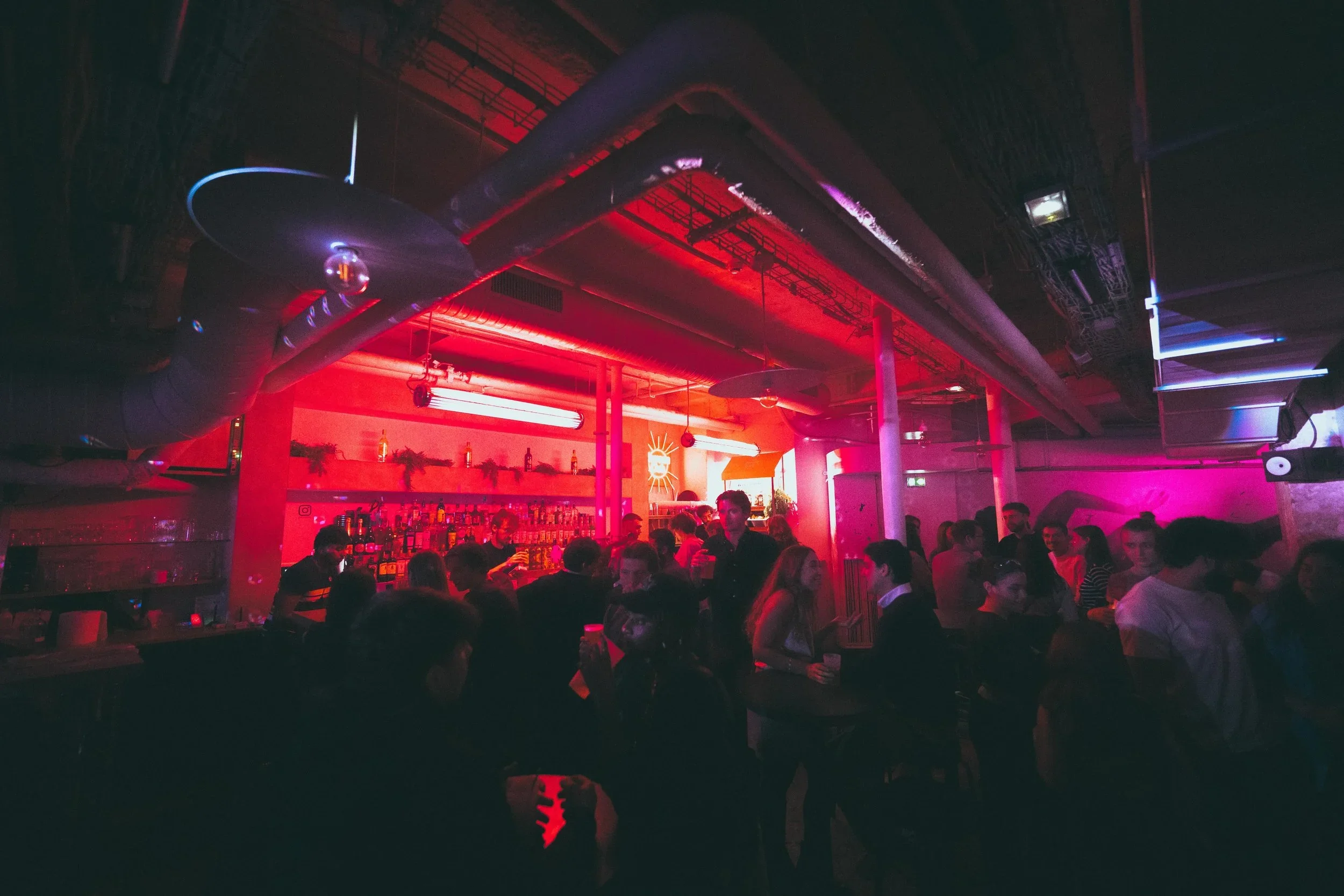 Une scène de bar ou de club avec des personnes discutant et buvant, ambiance lumineuse rose et violette, décor industriel avec tuyaux visibles au plafond.