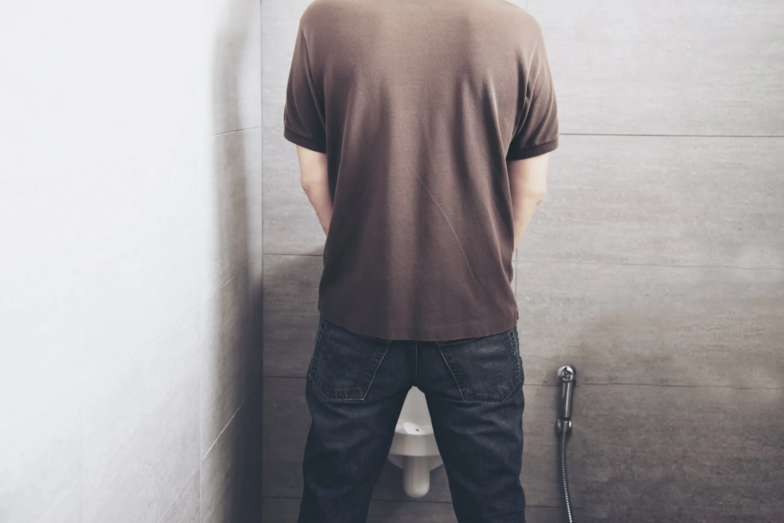 Person standing in front of a urinal in a bathroom with neutral-colored tiles, facing away from the camera.