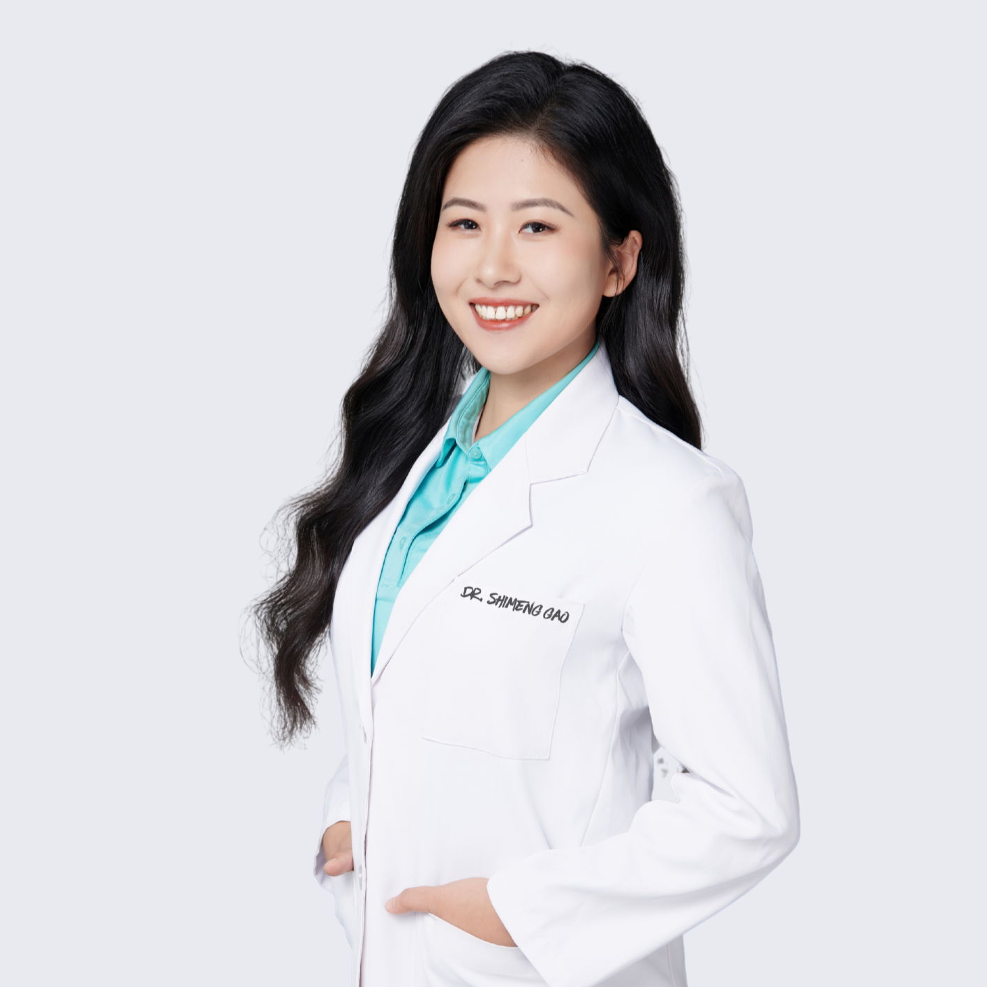 A portrait of a young female doctor of physical therapy(DPT) with long black hair smiling, wearing a white lab coat and a light blue shirt underneath, standing against a plain light background.