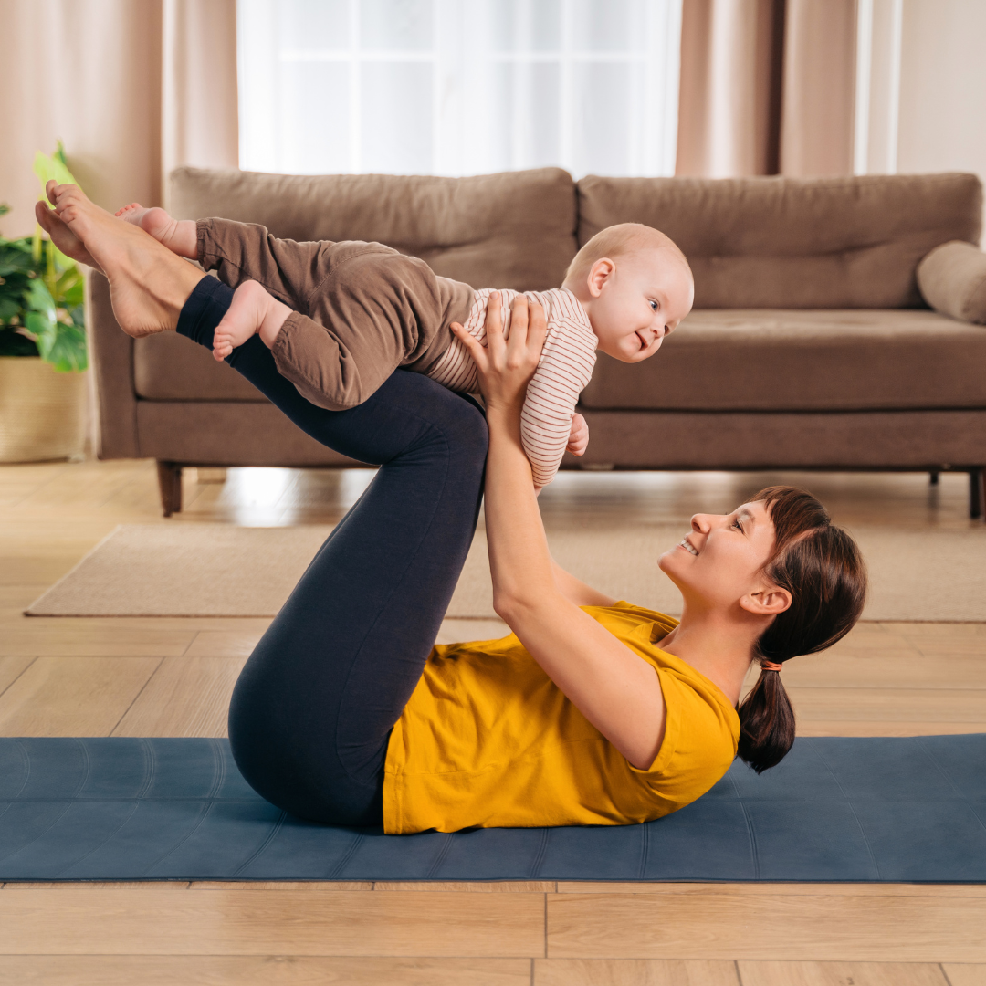 A woman is lying on a yoga mat doing a yoga pose while holding a baby above her on a living room floor with a brown couch and houseplant in the background.