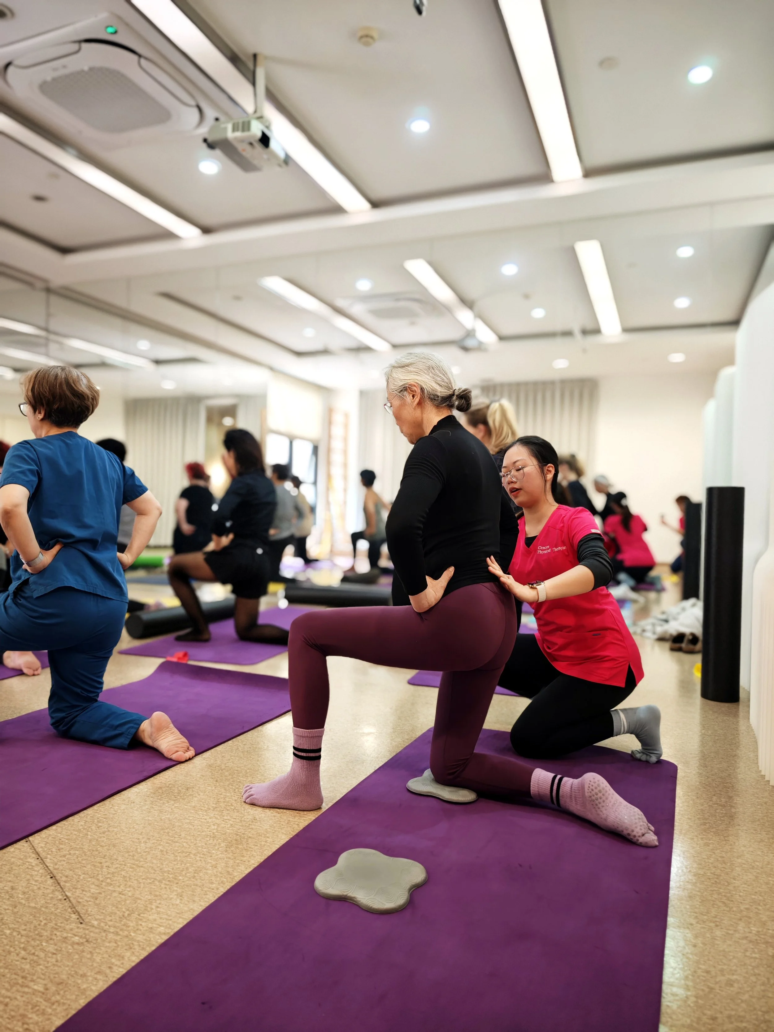 A group yoga class with people practicing on purple mats, with a trainer assisting a woman in a black top and purple pants in a yoga pose, in a well-lit room with ceiling lights and a projector.