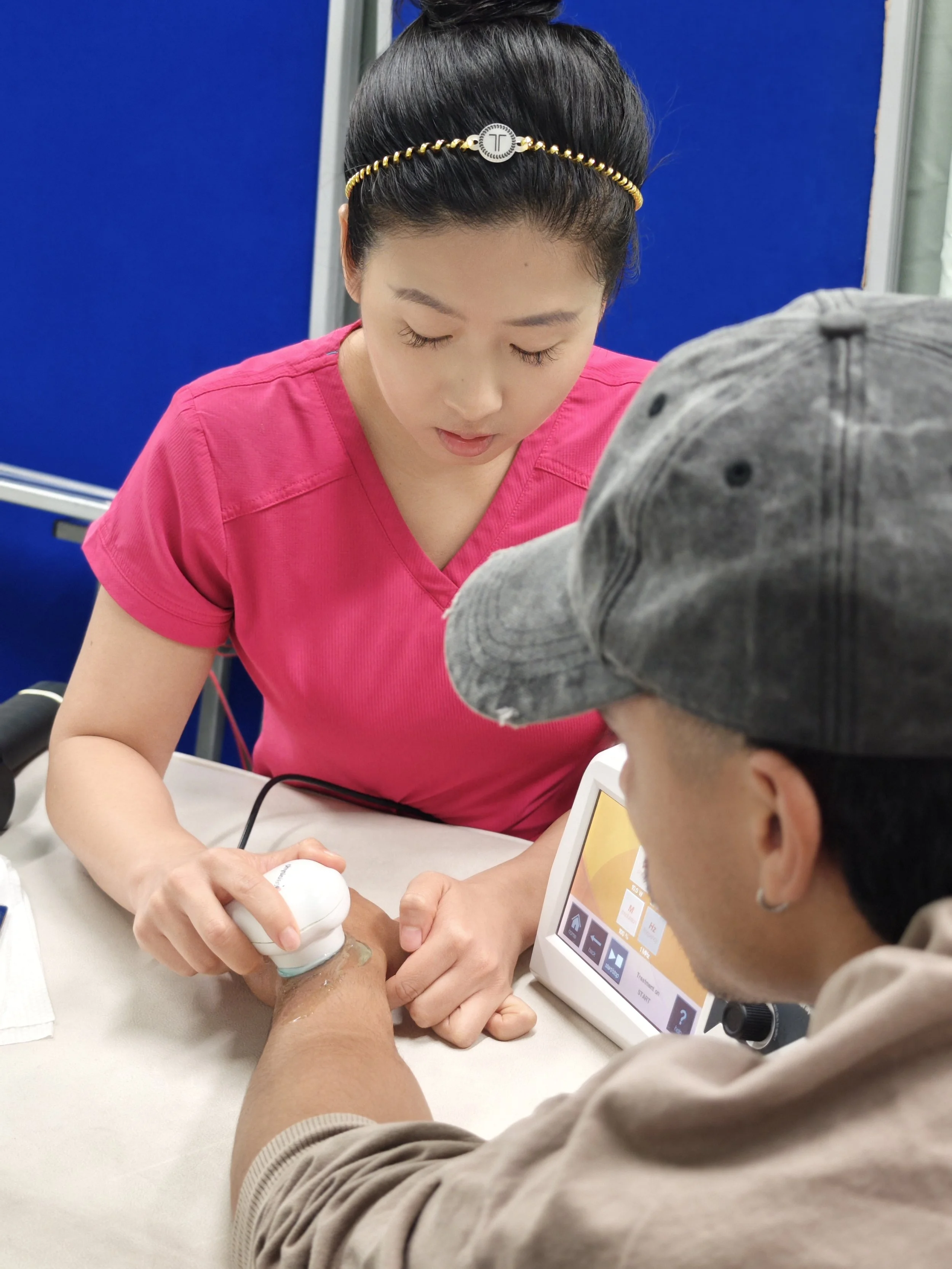 A woman in a pink shirt is conducting an ultrasound on a person's arm while a technician monitors the display screen.