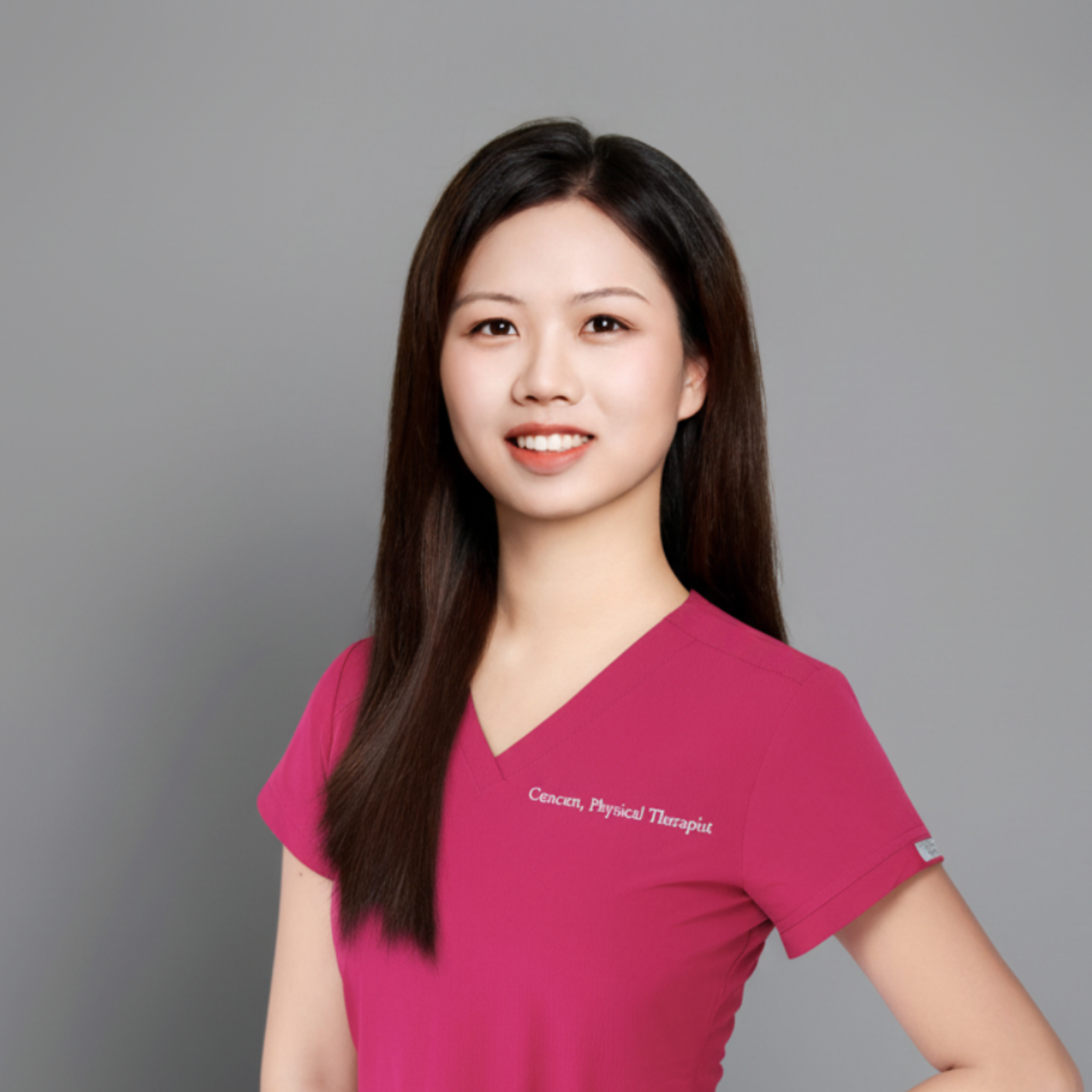 Portrait of a female physical therapist with long dark hair, wearing a pink medical scrub top with white embroidered text on the chest, standing against a gray background.