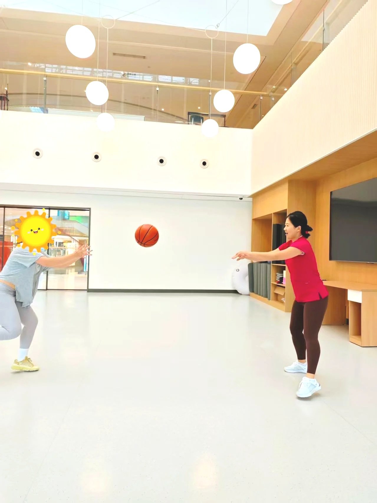 Two women playing basketball indoors with one woman shooting and the other reaching out to catch the ball, in a modern building with white and wooden walls and round ceiling lights.