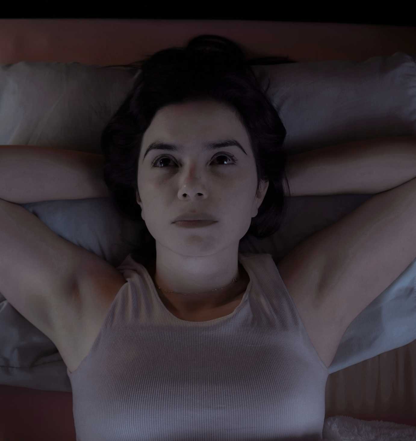 Woman lying in bed with her hands behind her head, looking up at the ceiling.