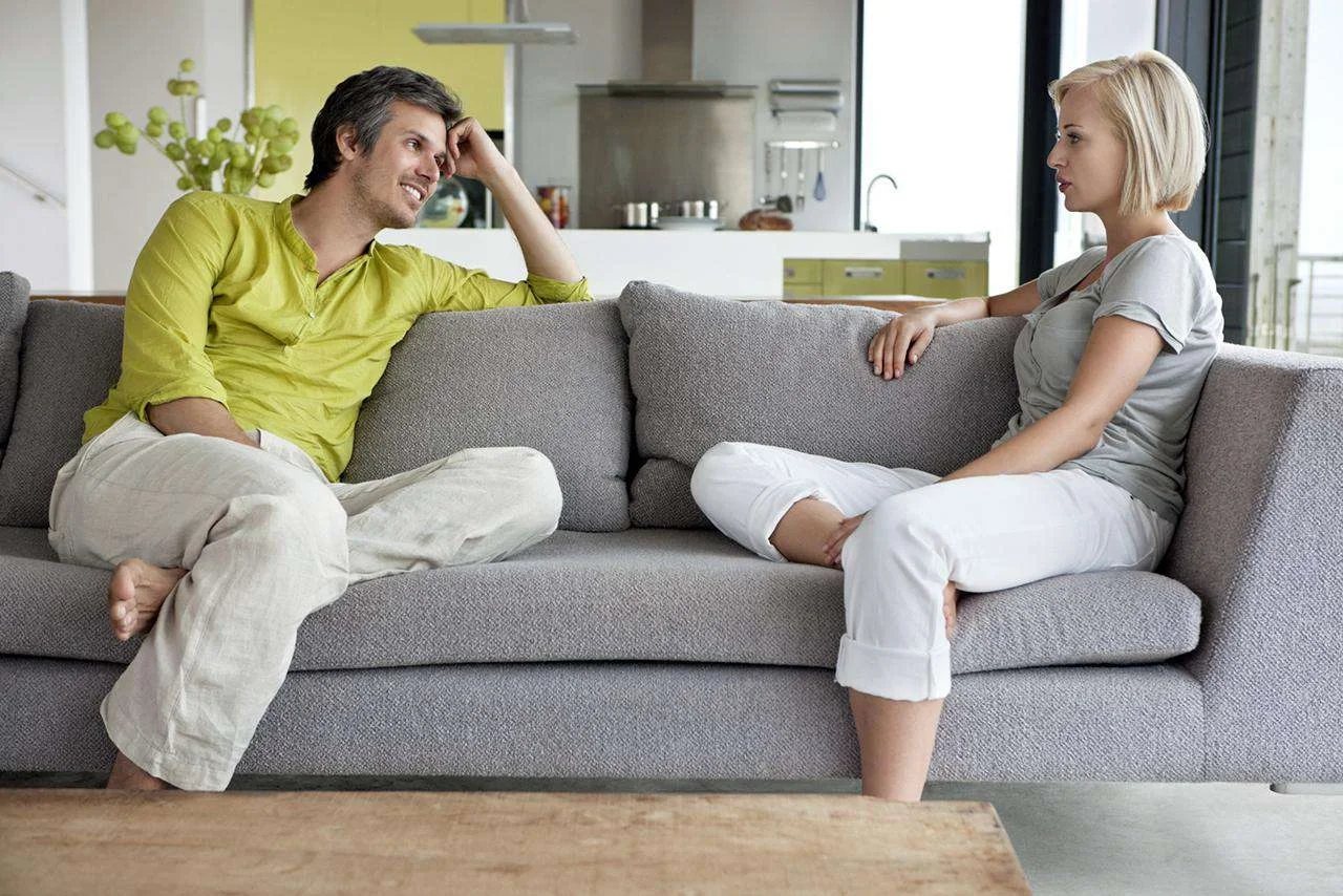 A couple sitting on a couch, smiling and talking together, symbolizing open communication and healthy conflict resolution.