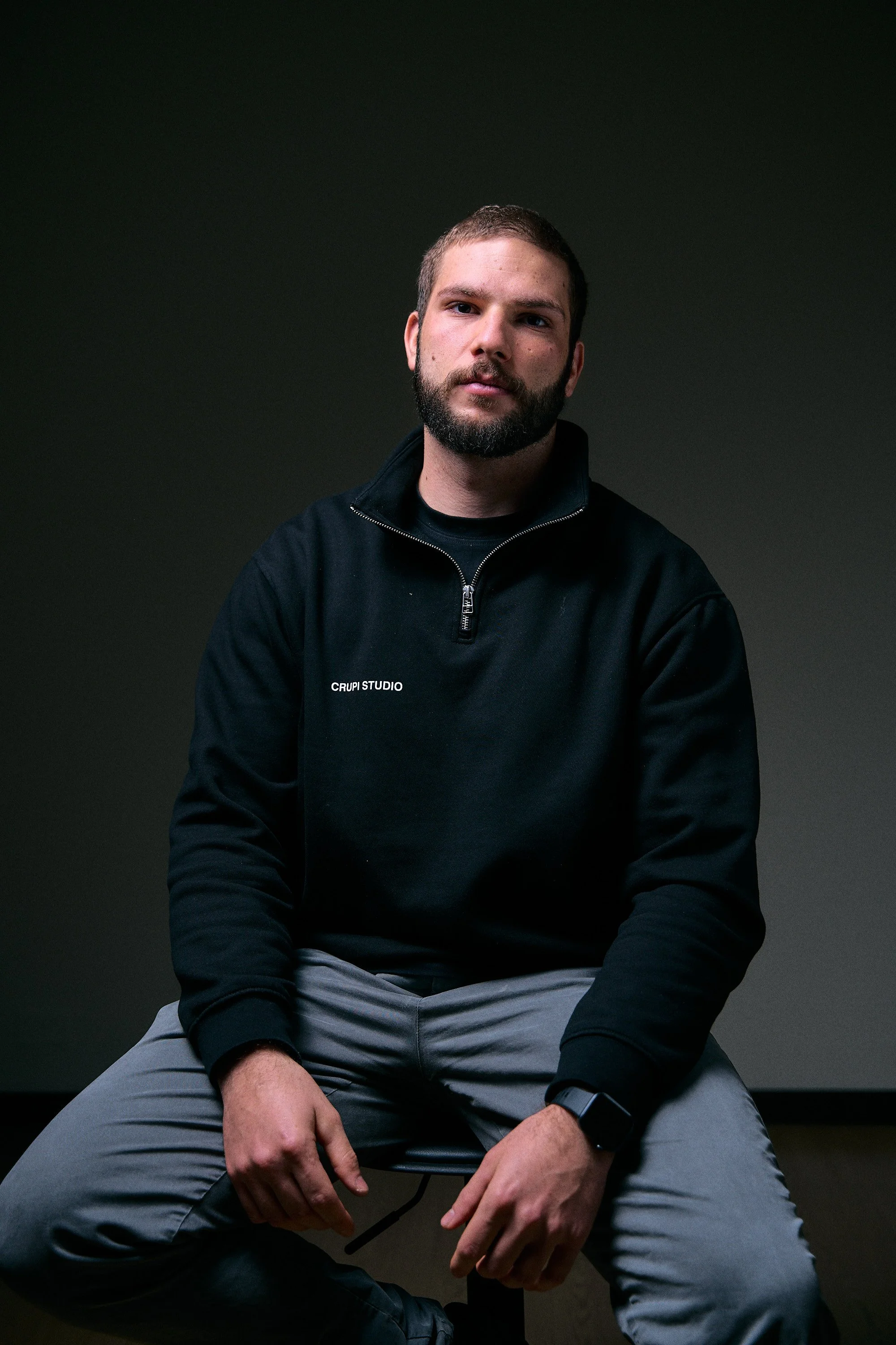 A young man with a beard and short hair, wearing a black T-shirt, sitting against a plain dark background.