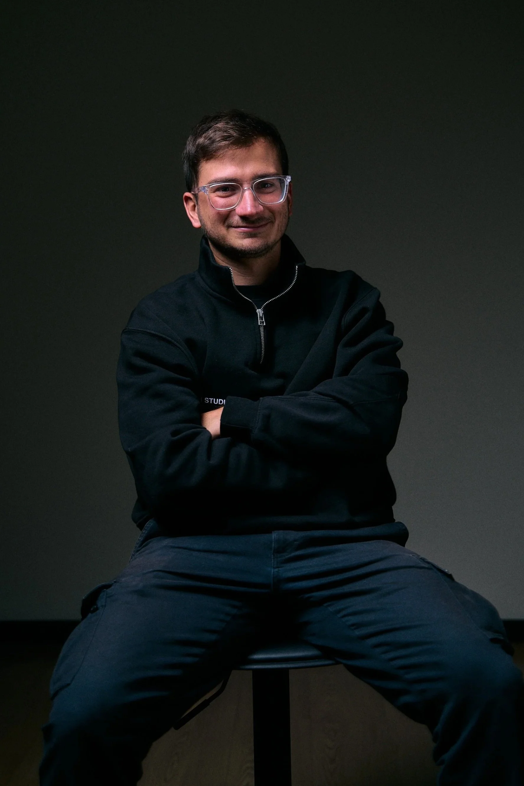 A young man with glasses, wearing a black shirt and blue jeans, sitting against a plain background.