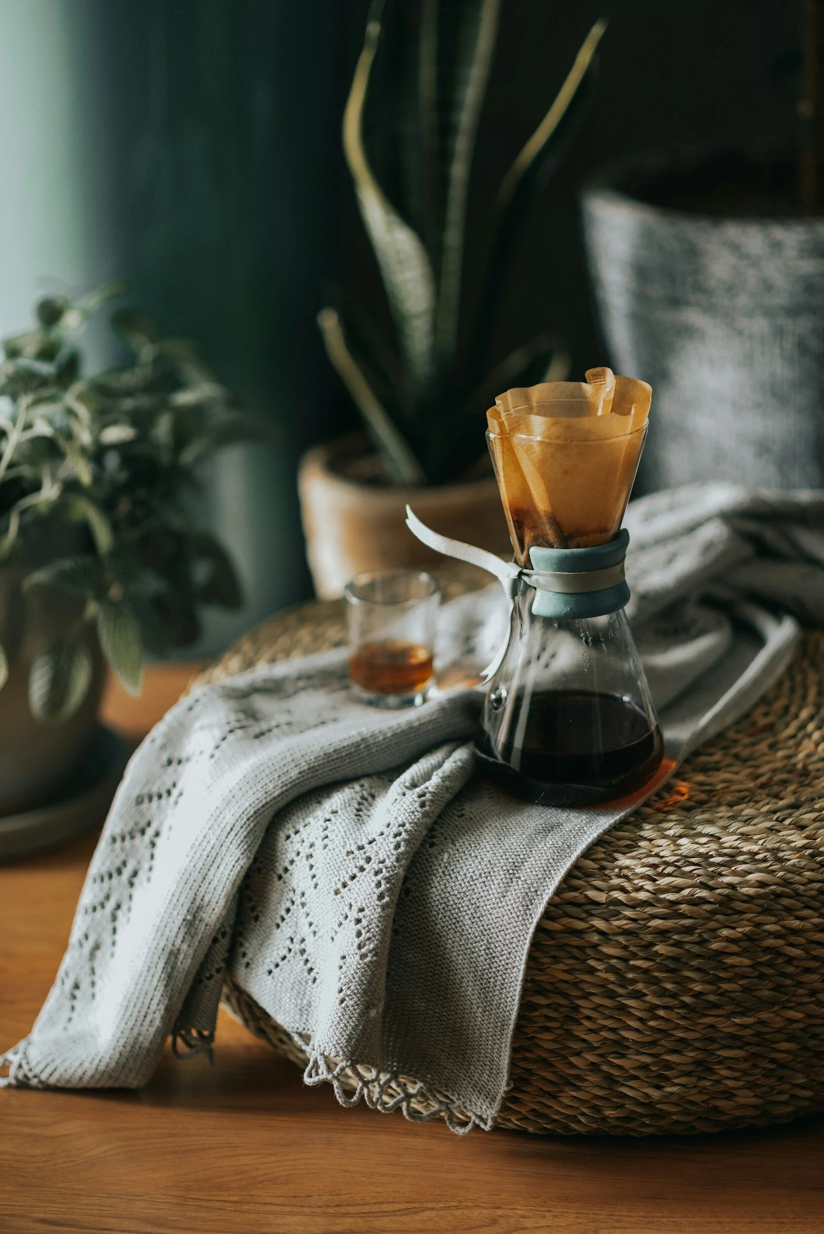 A Chemex coffee maker filled with dark coffee sits on a wicker basket, with a cloth napkin draped over it. A small glass of coffee is nearby, and there are potted plants in the background.
