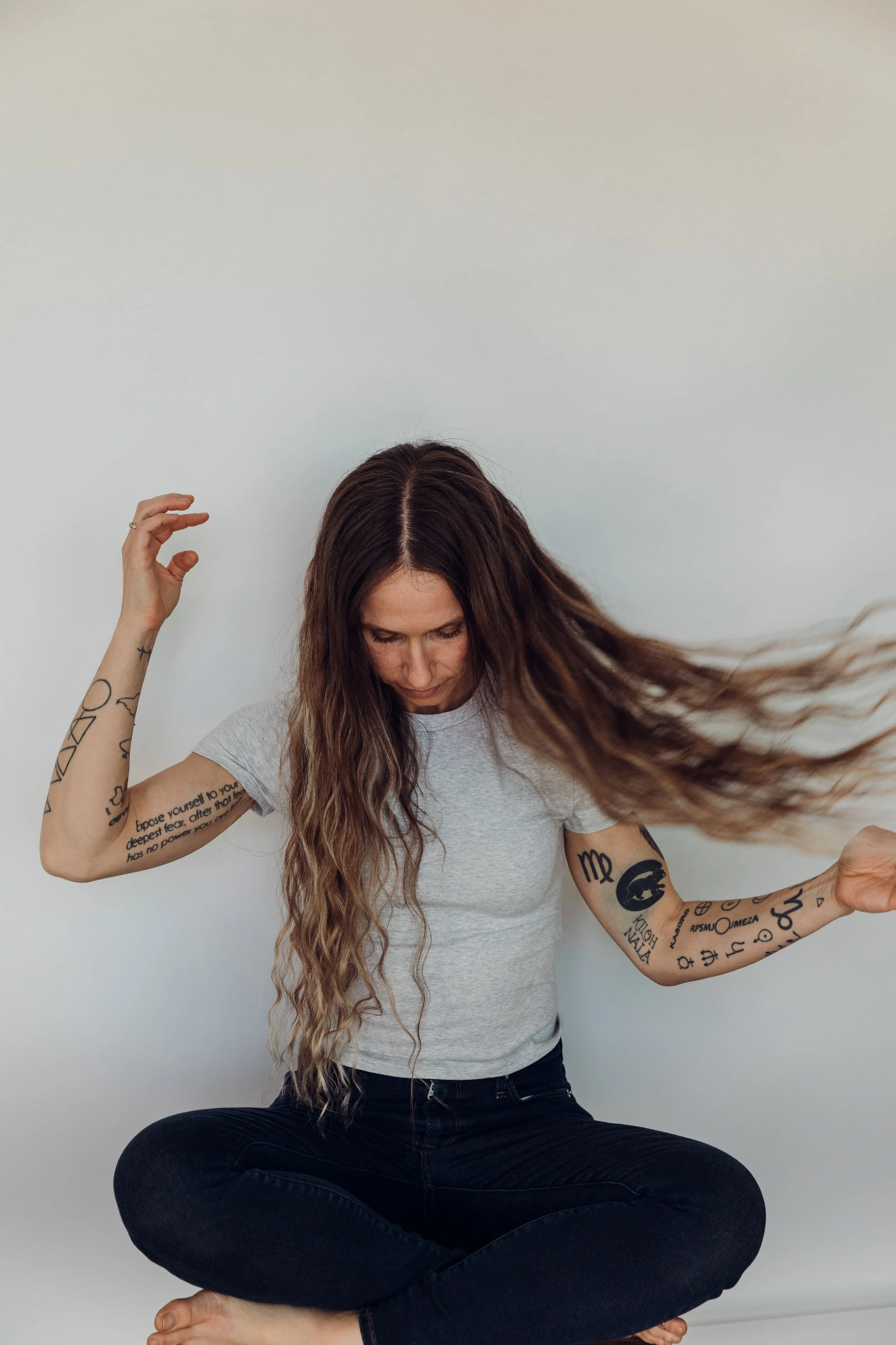 Woman with long, wavy hair sitting cross-legged against a plain white wall, with tattoos on her arms, wearing a gray t-shirt and black jeans, flipping her hair to the side.