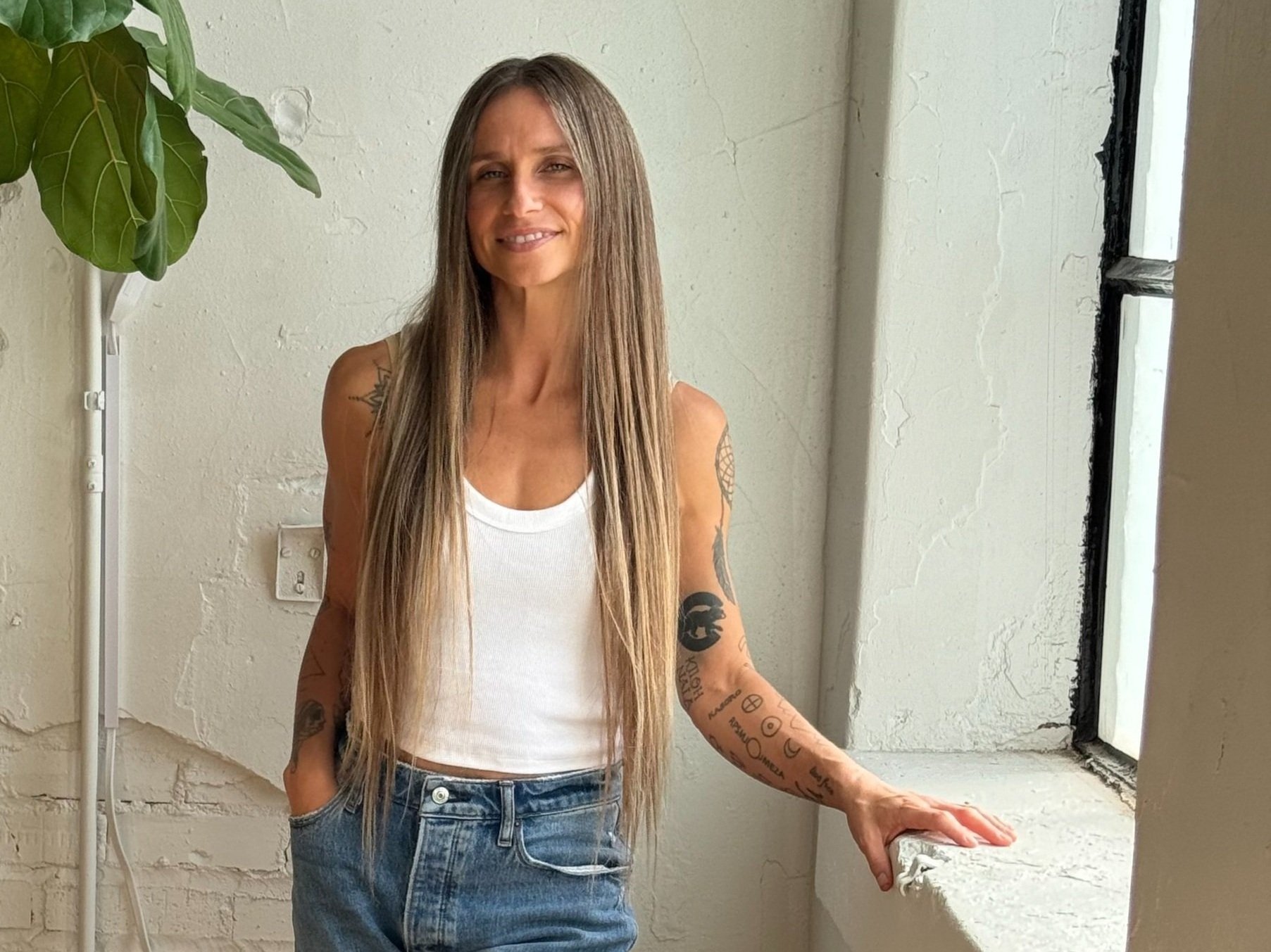 A person with long straight hair wearing a white tank top and blue jeans stands indoors by a window. They have tattoos on their arms and are next to a green potted plant.