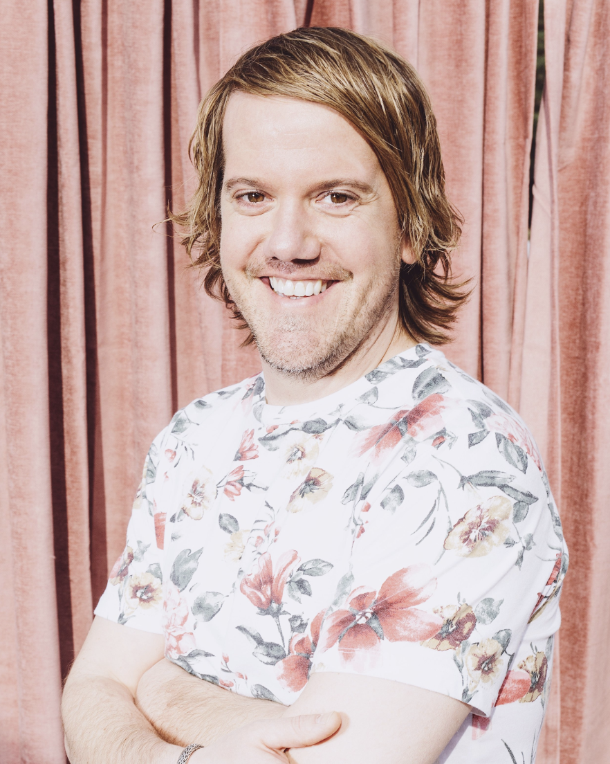 Smiling person in floral shirt with arms crossed, standing in front of pink curtains.