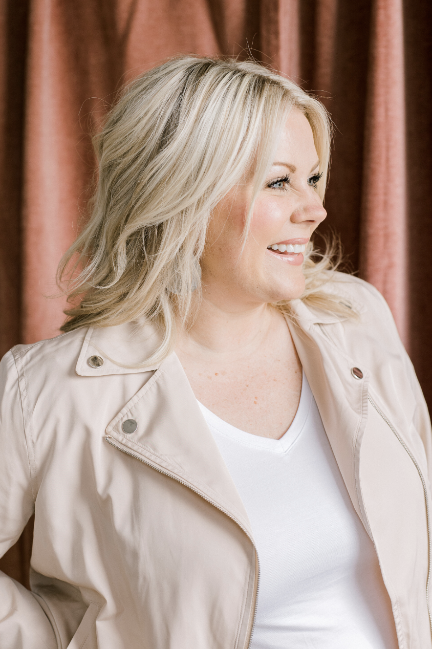 Smiling woman with blonde hair wearing a beige jacket and white shirt, looking to the side with a brown curtain backdrop.