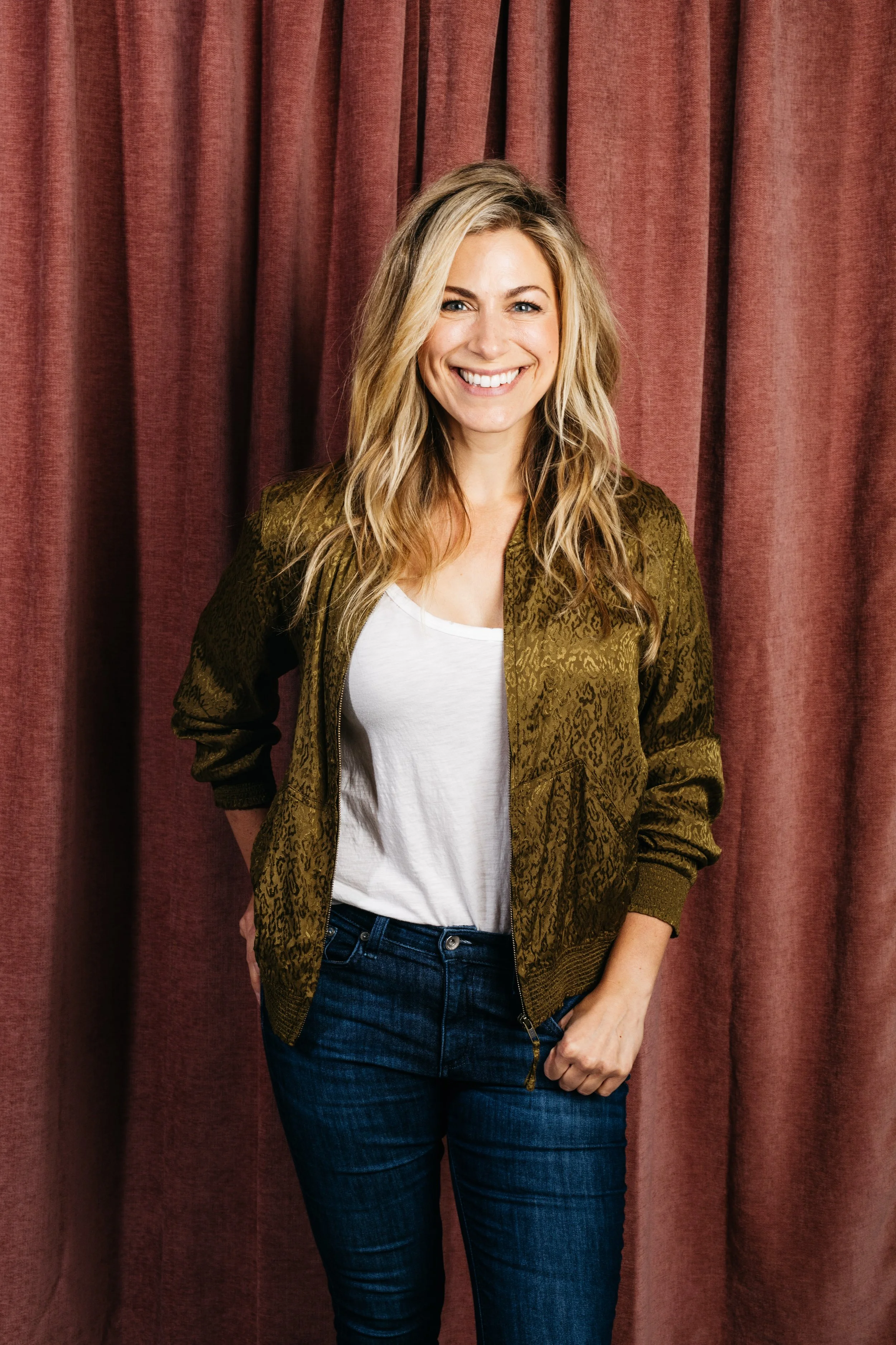 Smiling woman with long blonde hair, wearing a green jacket and jeans, standing in front of red curtains.