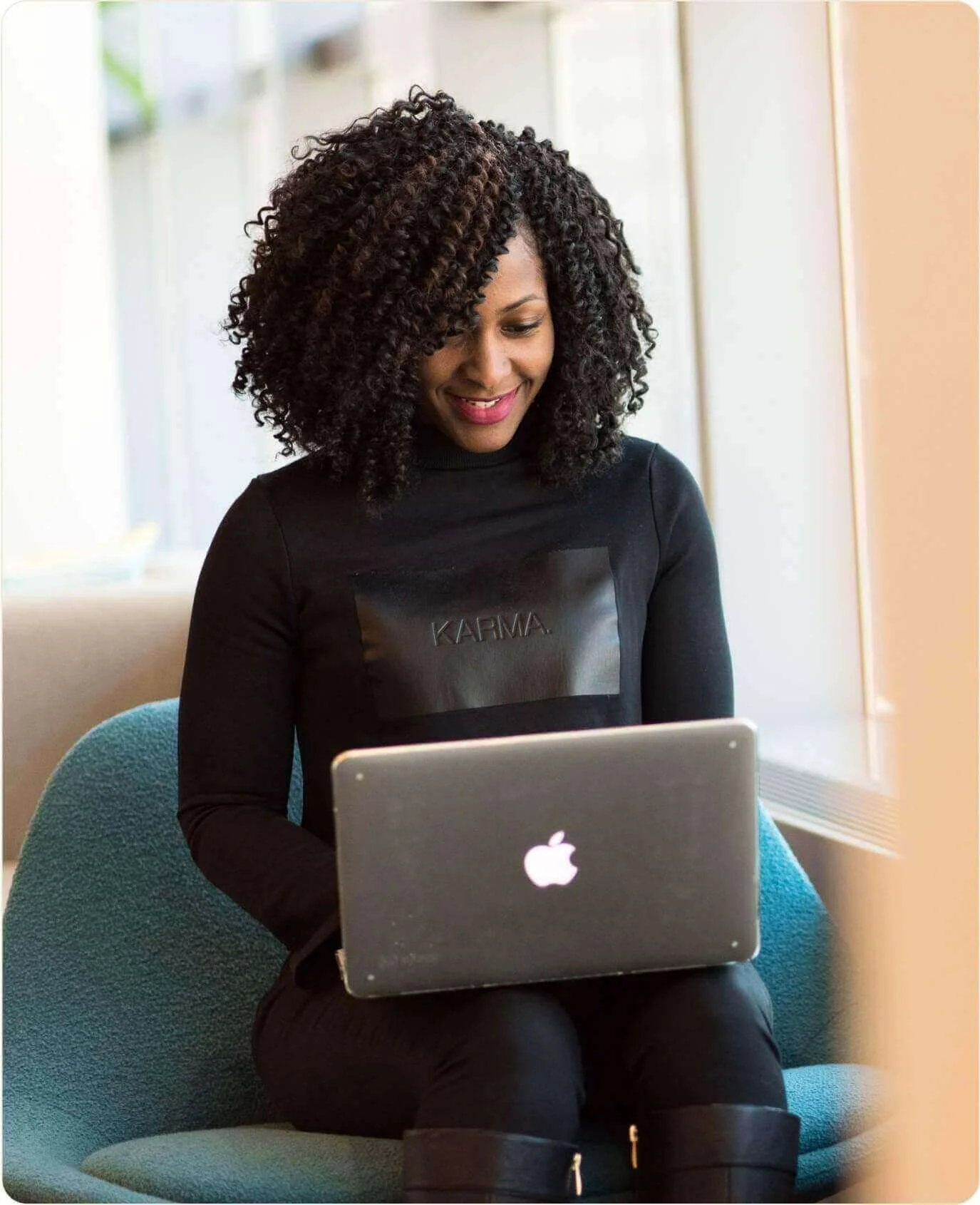 A woman with curly dark hair smiling while sitting in a blue chair and working on a silver MacBook laptop with an illuminated Apple logo.