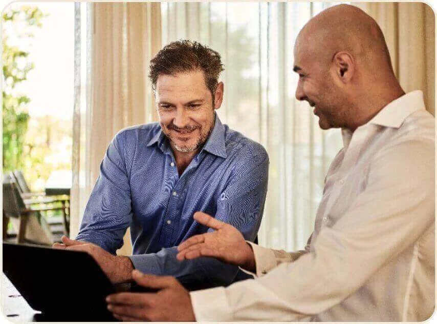 Two men sitting at a table looking at a laptop, with one man smiling and pointing at the screen.