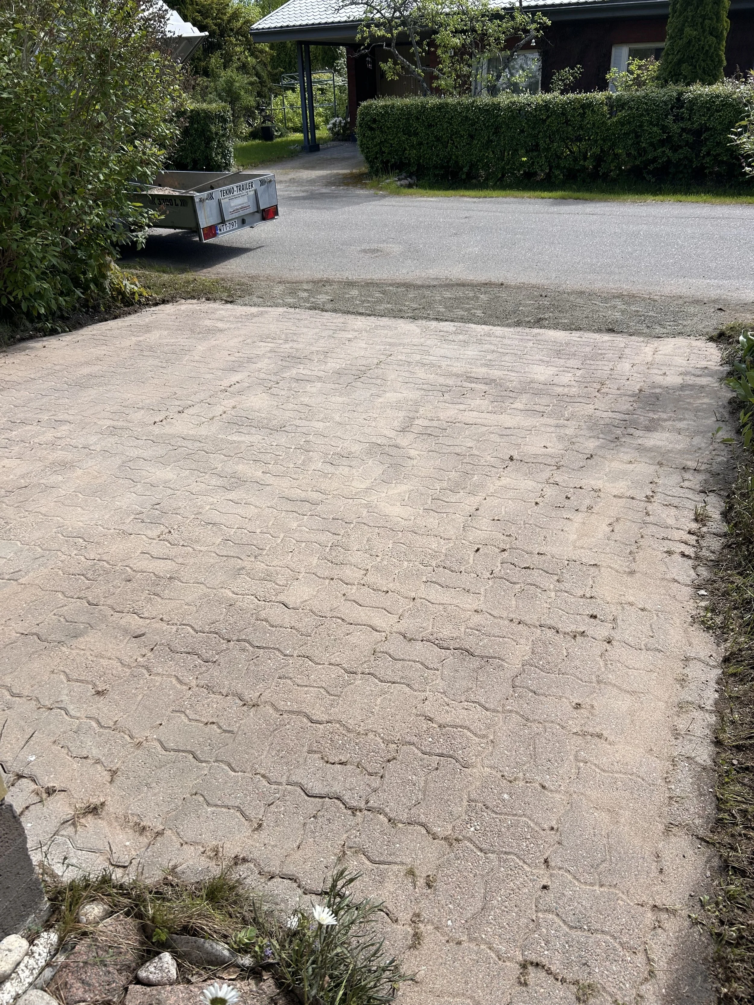 Pihatie ja piha-alueen osa, nurmikkoa, pensaita, kukkapenkki ja talo taustalla.