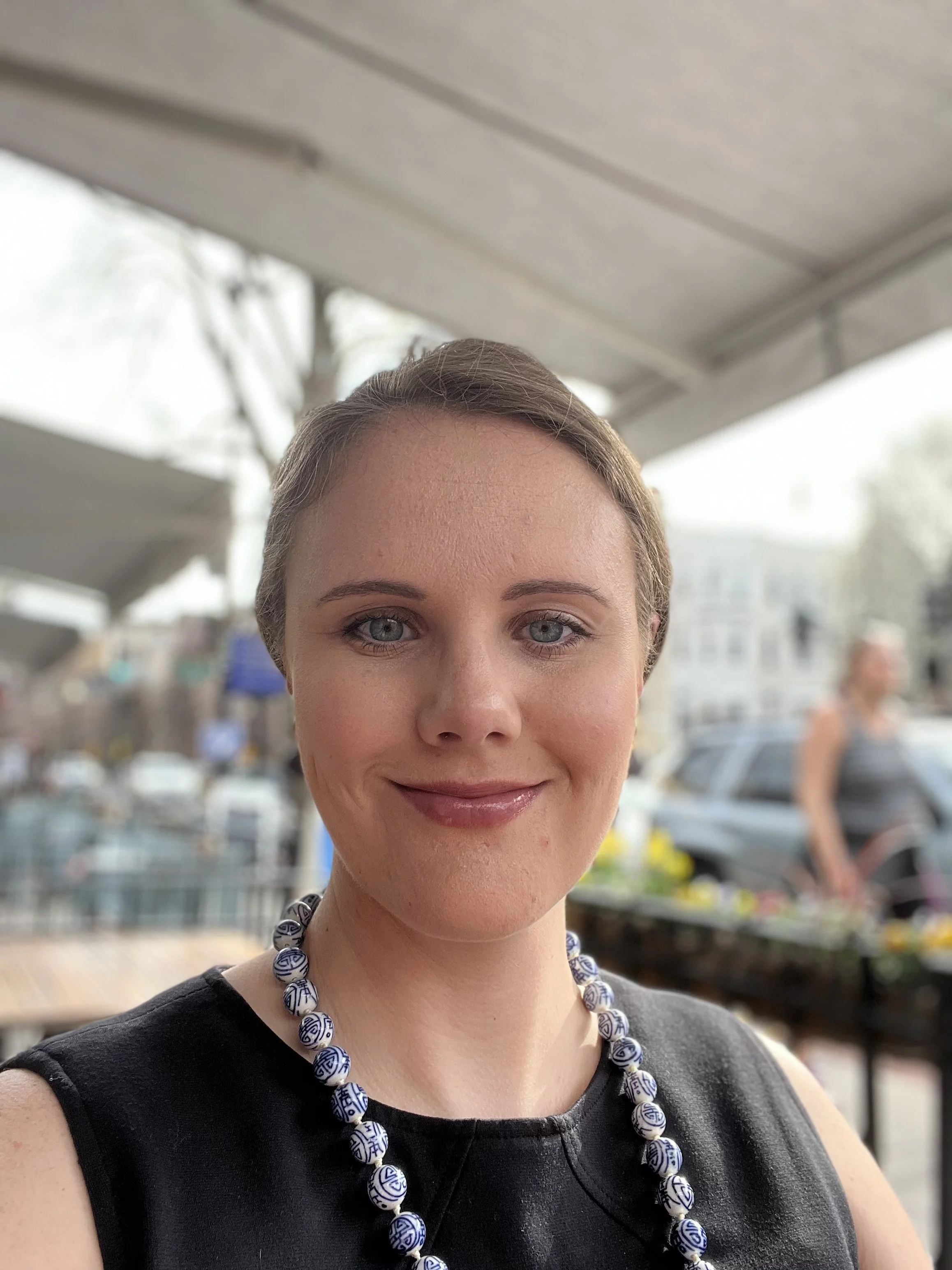 Person smiling, wearing a blue and white beaded necklace, in an outdoor setting with blurred background of people and buildings.