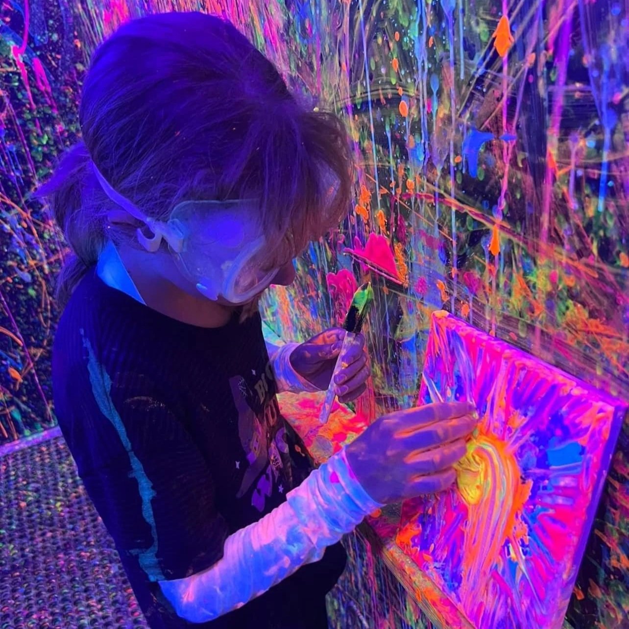 Person with safety glasses and gloves creating neon-colored drip art on canvas in a blacklight room.