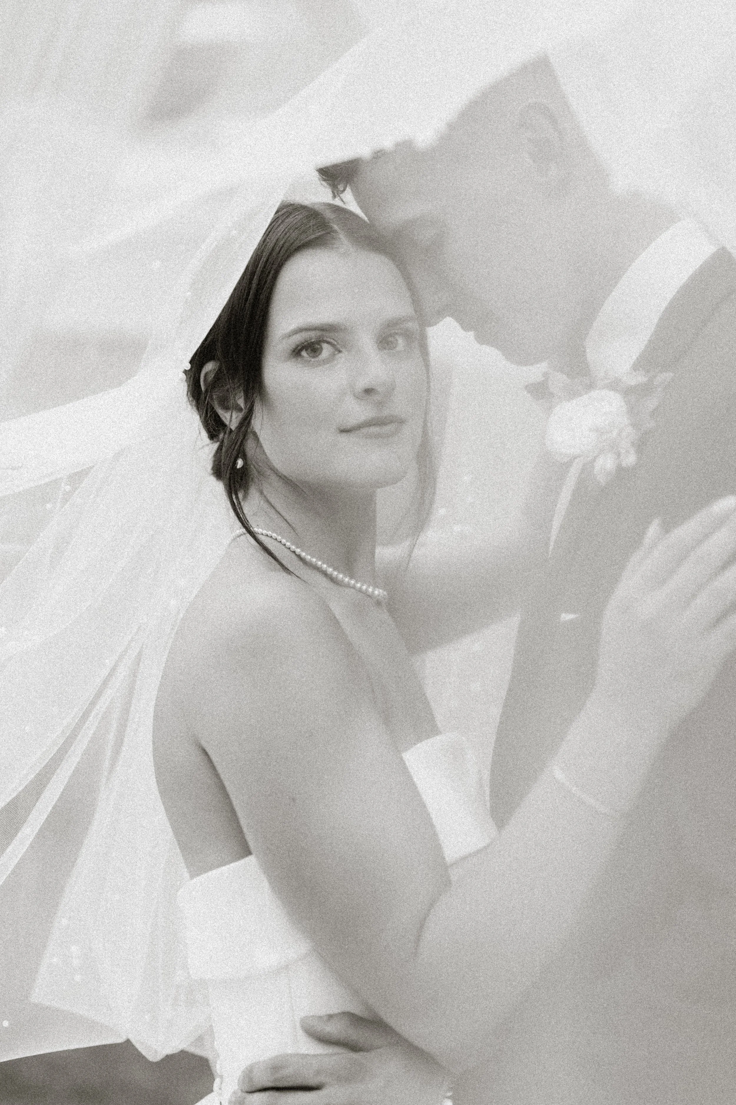 Black and white photo of a bride and groom embracing, with the groom leaning over and the bride looking at the camera, wearing a wedding dress and veil.