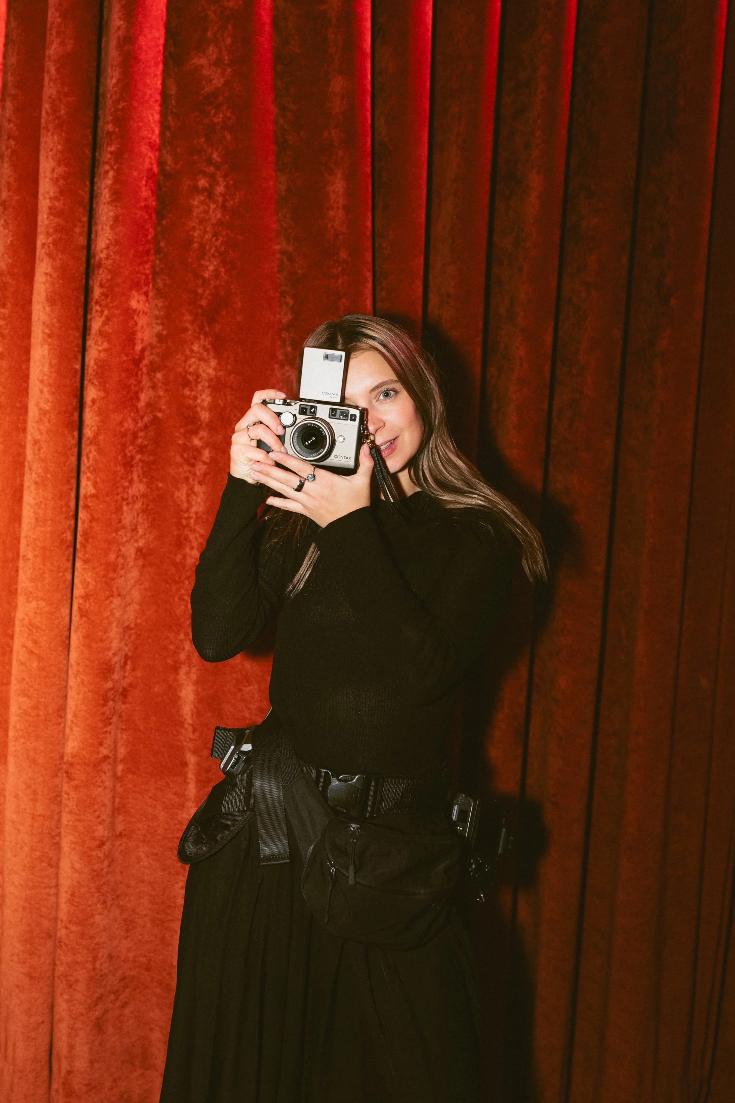 Young woman taking a photo with a vintage camera in front of red velvet curtains.