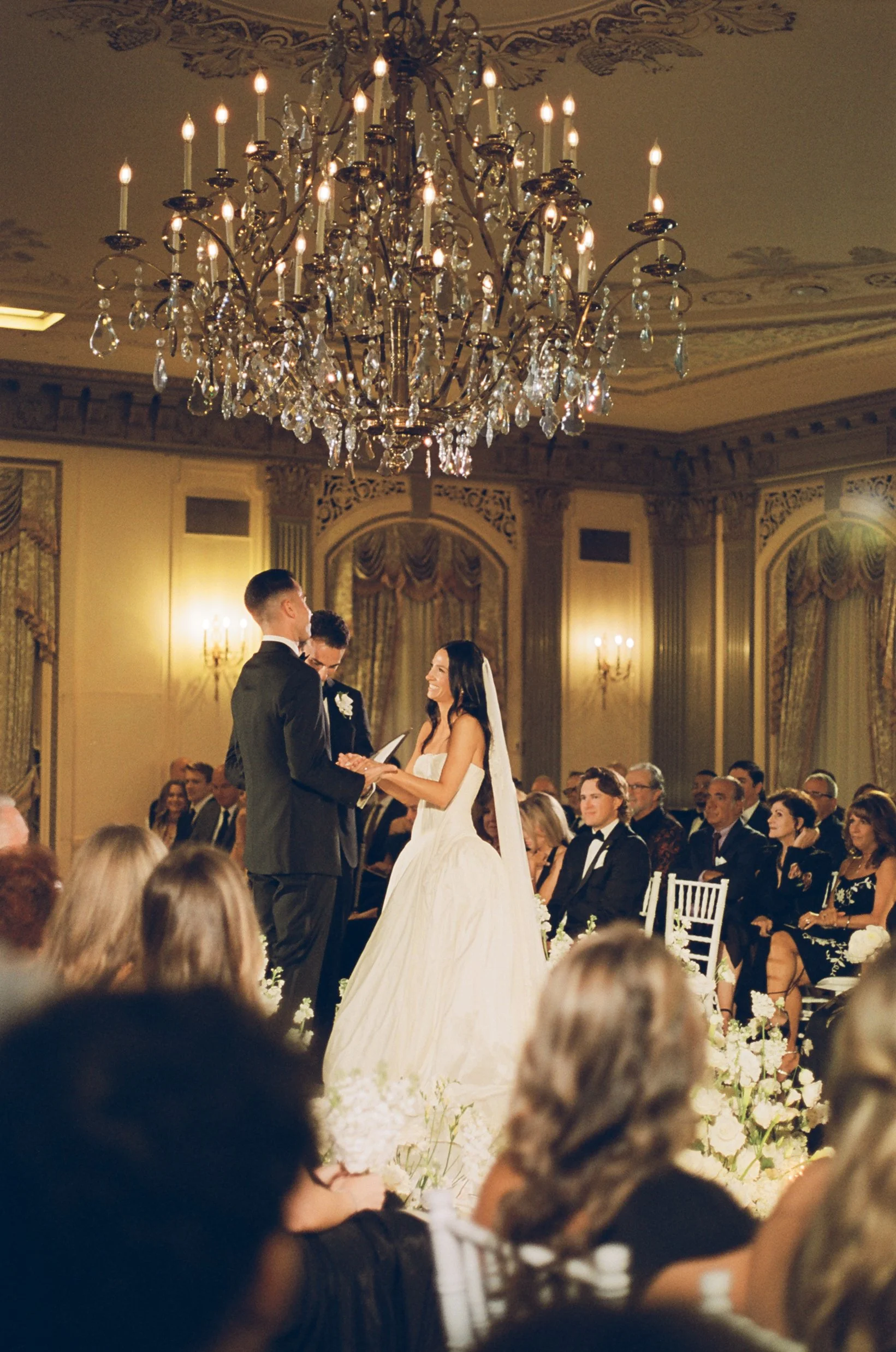 A wedding ceremony inside a grand, elegant hall with a chandelier hanging from the ceiling, a bride and groom exchanging vows in front of seated guests.