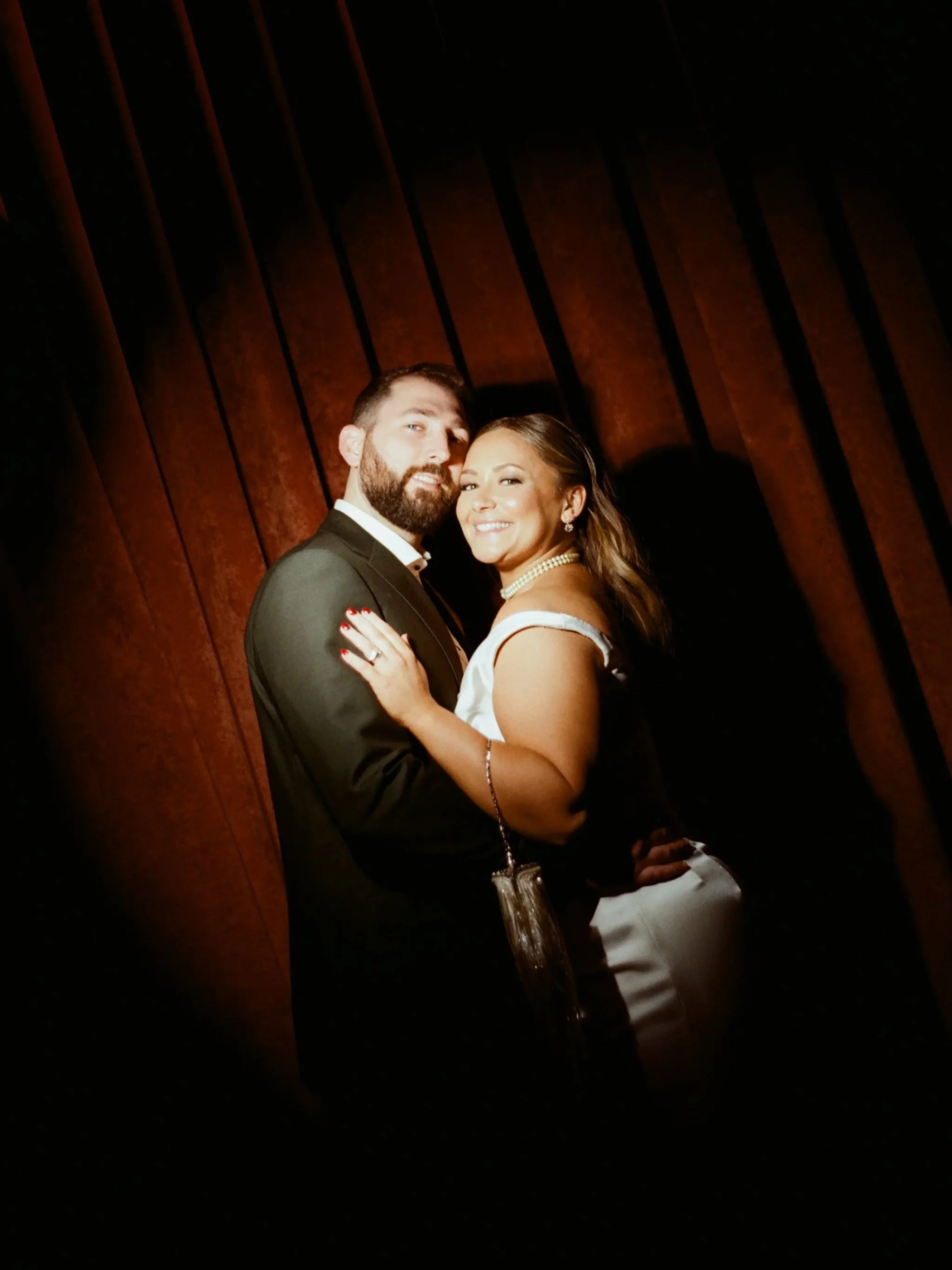 A man and woman dressed in formal attire pose together in front of a wooden wall, smiling at the camera.