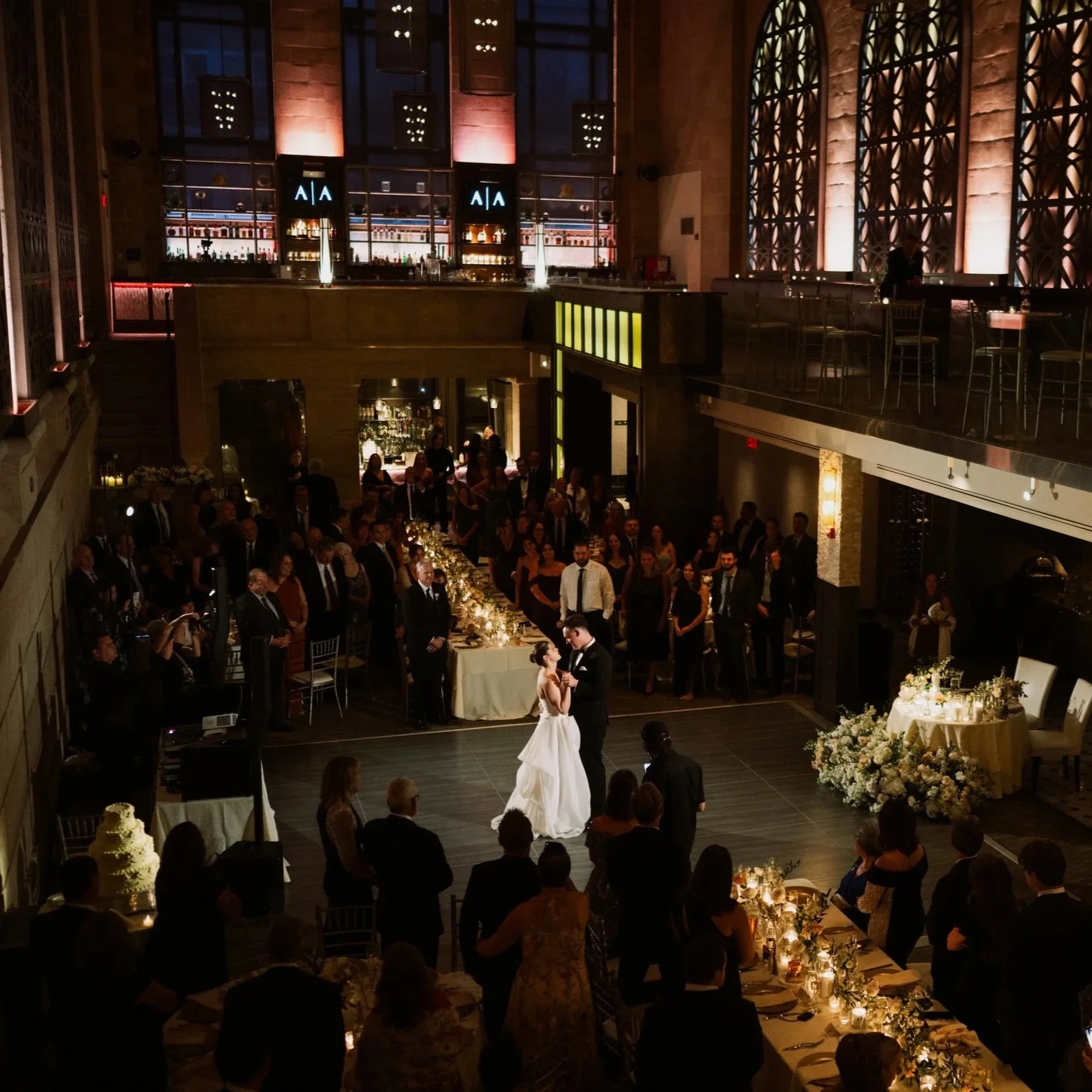 A bride and groom dancing at their wedding reception in an elegant, dimly lit venue with many guests watching. The bride is in a white gown, and the groom is in a black tuxedo. There are decorated tables, floral arrangements, and a cake visible in the scene.
