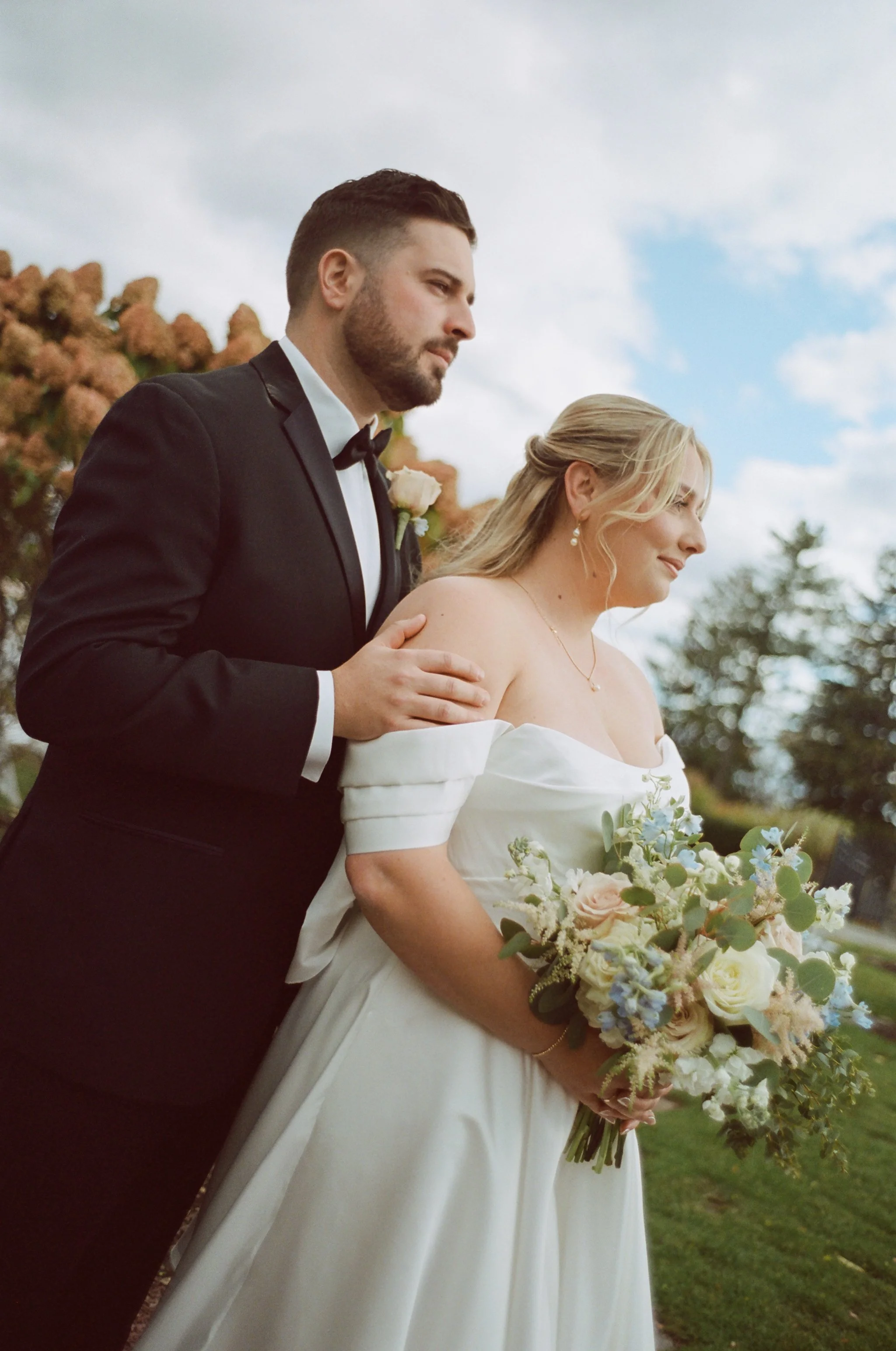 A bride and groom standing outdoors, with the groom gently touching the bride's shoulder, both dressed in wedding attire.