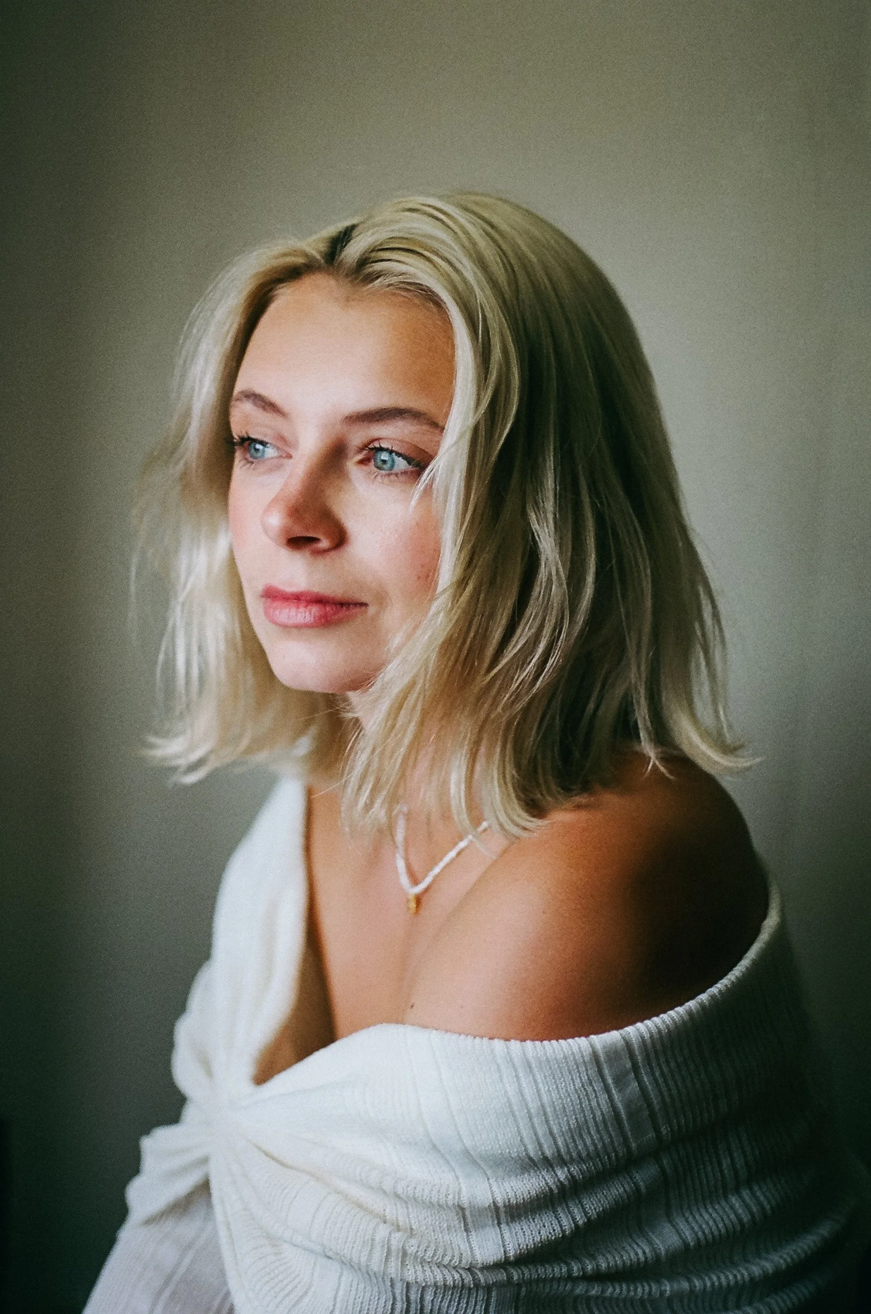 A woman with shoulder-length blonde hair, blue eyes, and fair skin, wearing a white off-the-shoulder sweater and a delicate necklace, poses against a neutral background.