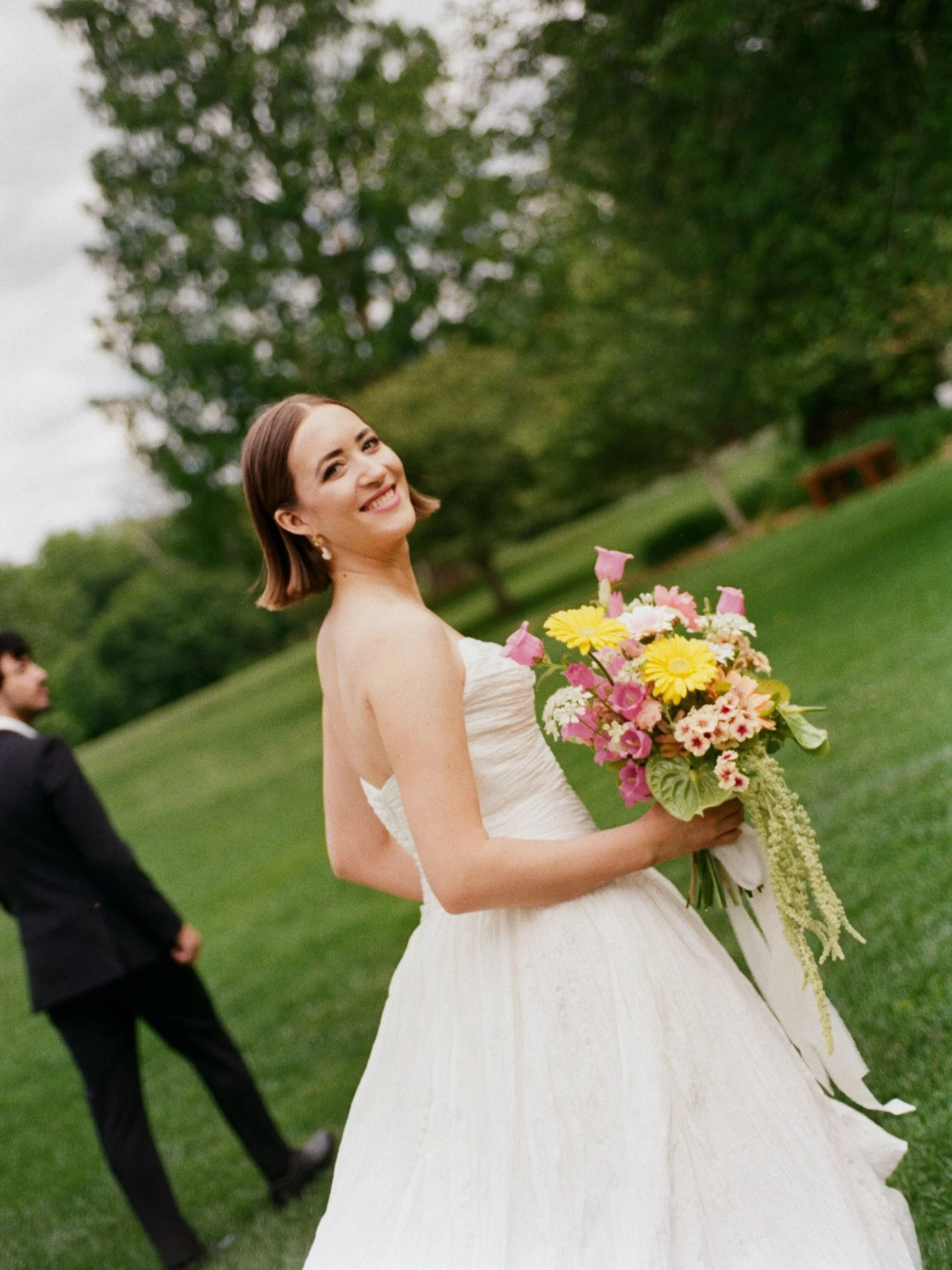 A smiling bride in a white wedding dress holding a colorful bouquet outdoors in a park with green grass and trees.