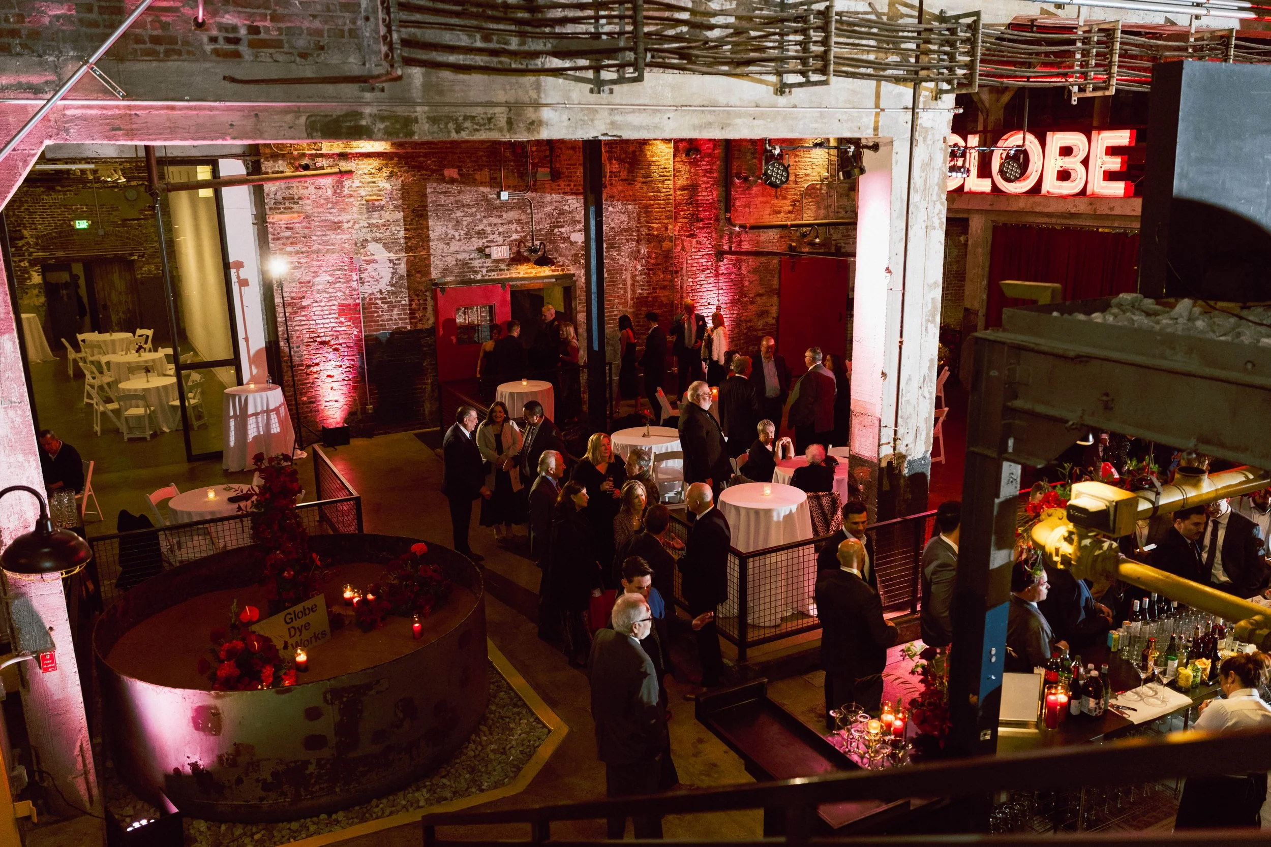 Indoor event with guests socializing around decorated cocktail tables, bar area with bartenders, exposed brick walls, and neon sign that reads 'GLOBE' in a warehouse-style venue with industrial decor.