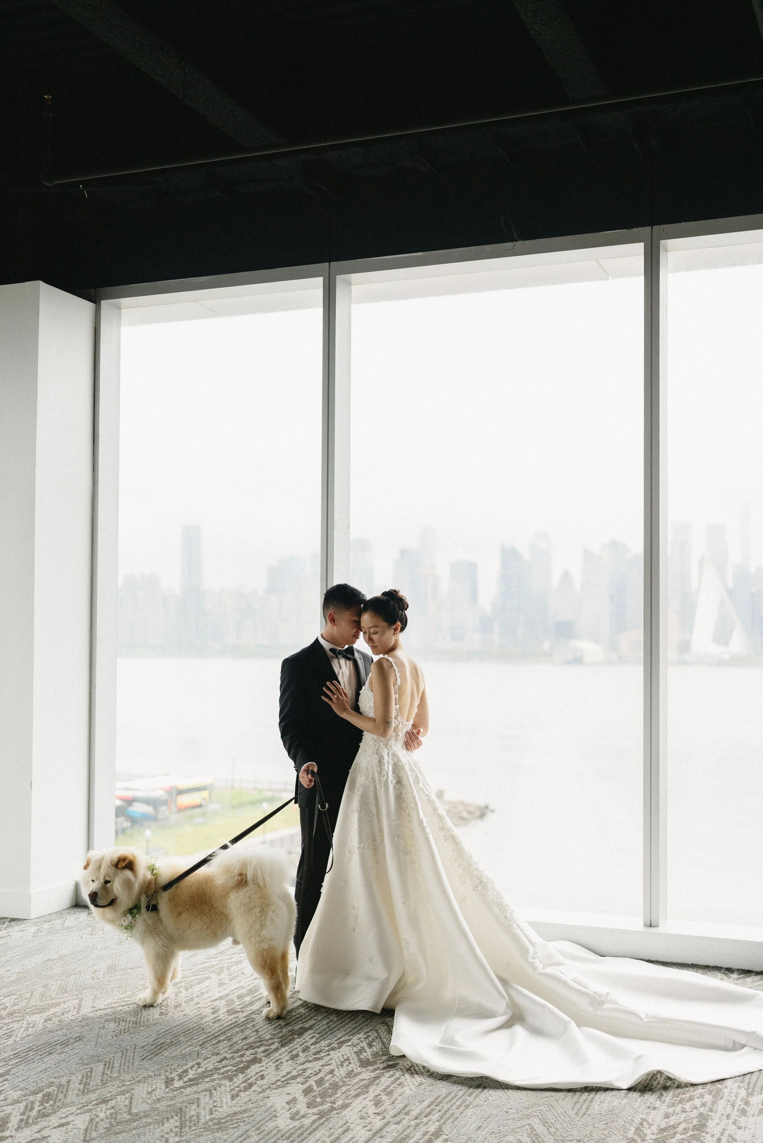 A bride and groom standing close together near a large window with a city skyline in the background, with the groom holding a fluffy dog.