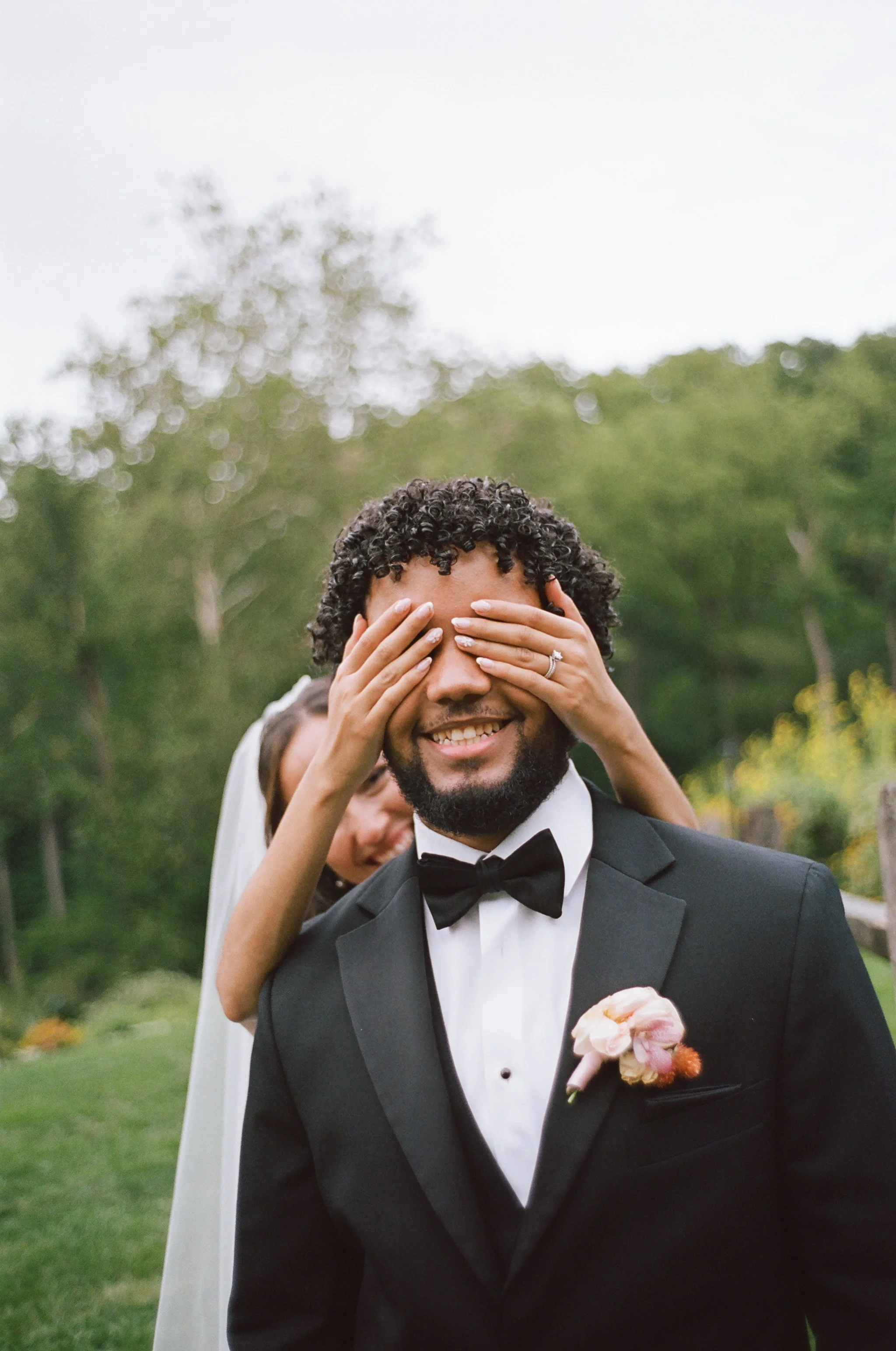 A groom in a black tuxedo and bow tie smiling as a woman covers his eyes with her hands, outdoors with trees in the background.