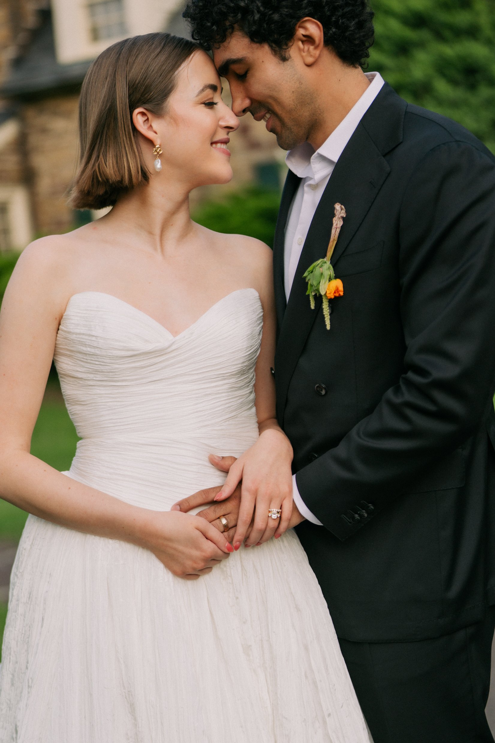 A bride and groom with foreheads touching, smiling, holding hands, outdoors, on a wedding day.