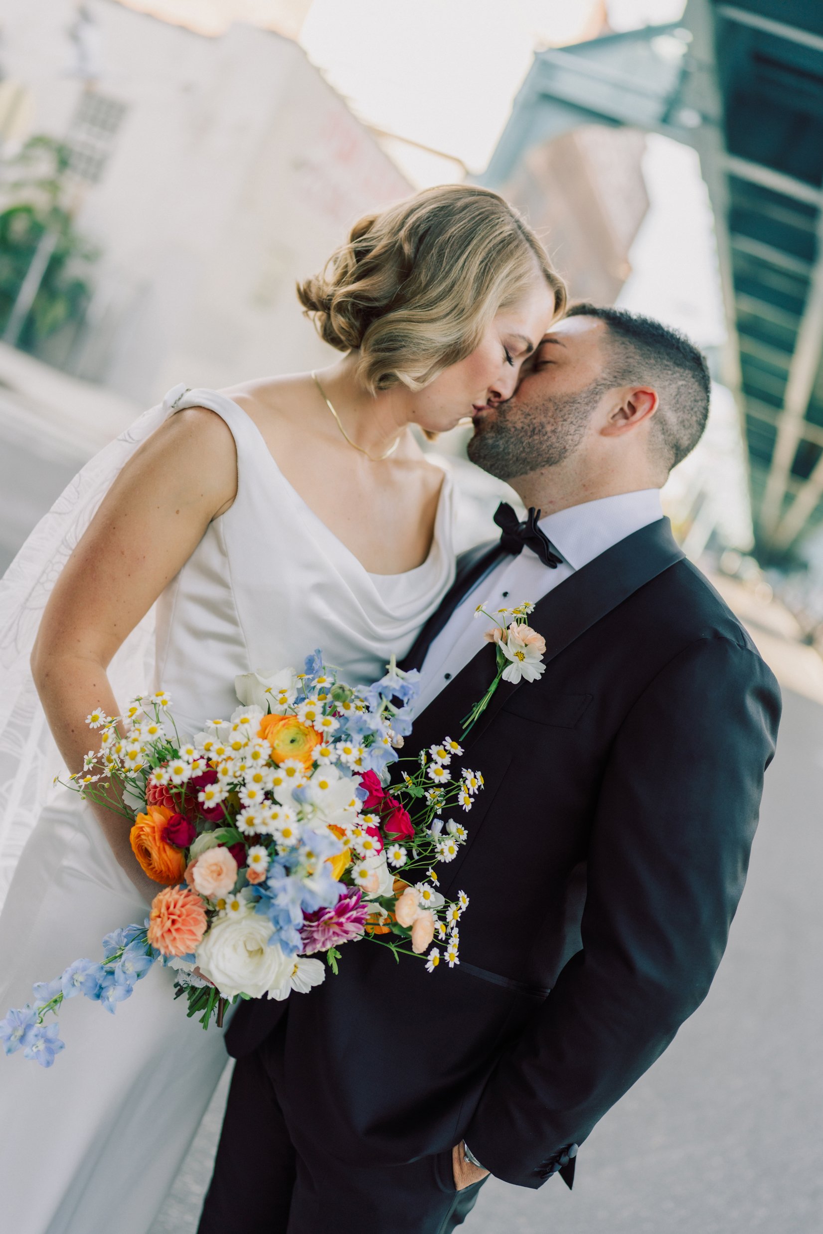 A bride and groom kiss on a city street, with the bride holding a vibrant bouquet of colorful flowers, during a wedding photoshoot.