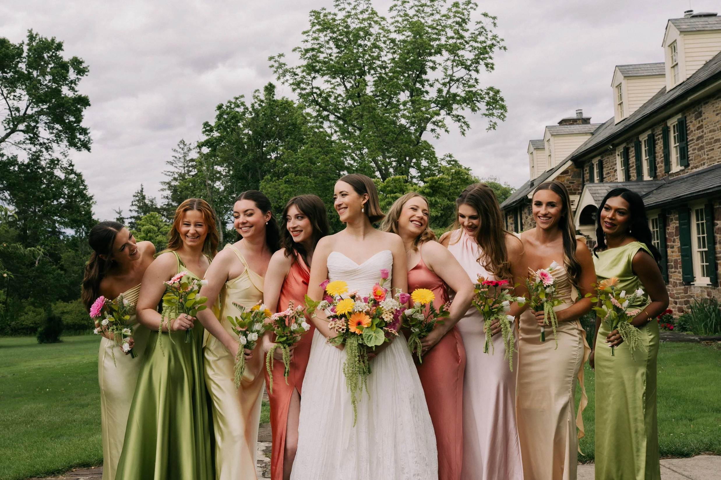 A bride in a wedding dress with nine bridesmaids in pastel-colored dresses, all holding bouquets, outside in front of a large stone house with trees and cloudy sky in the background.