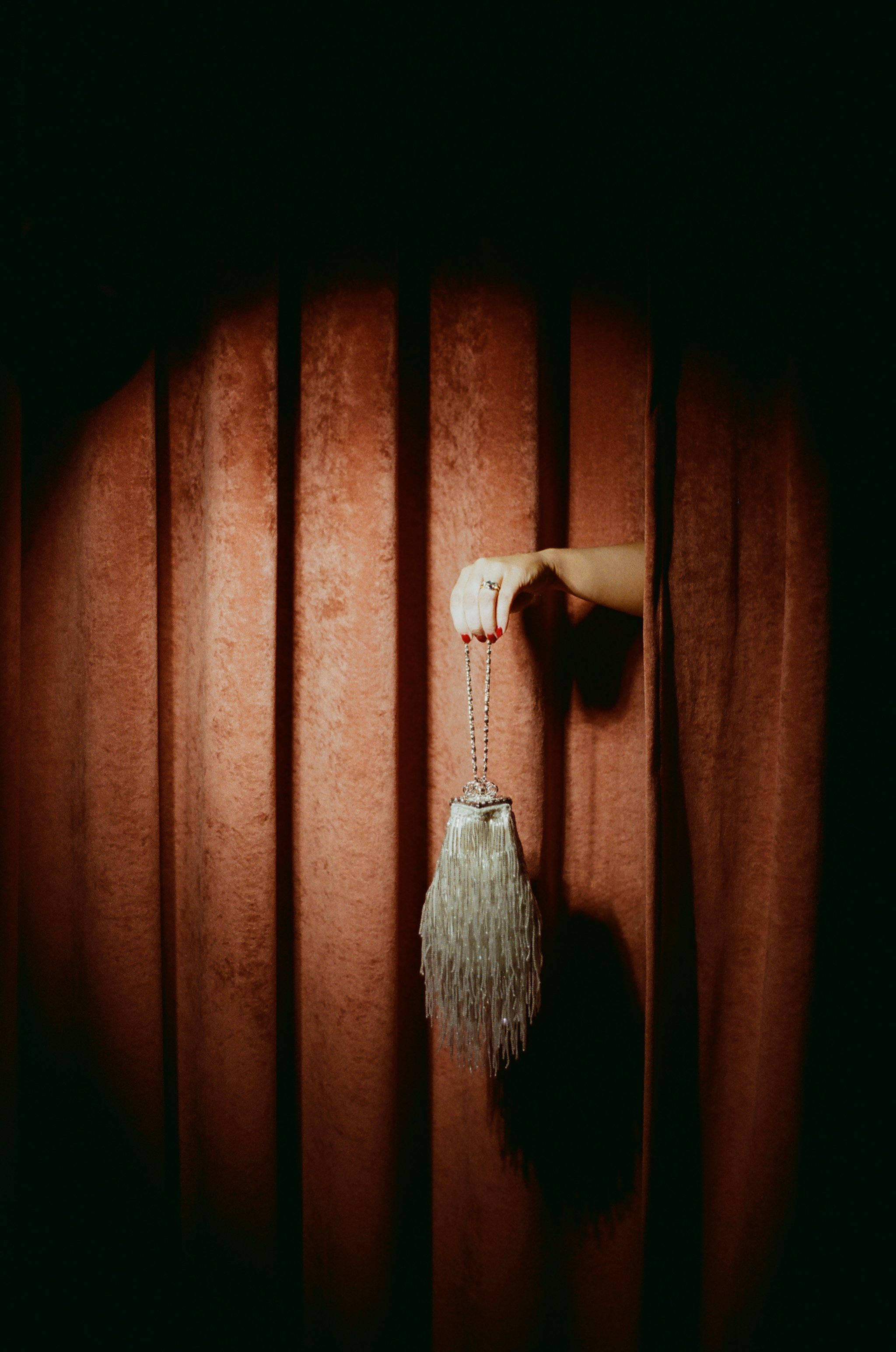 A person's hand with red nail polish holding a silver tassel purse against a red curtain.