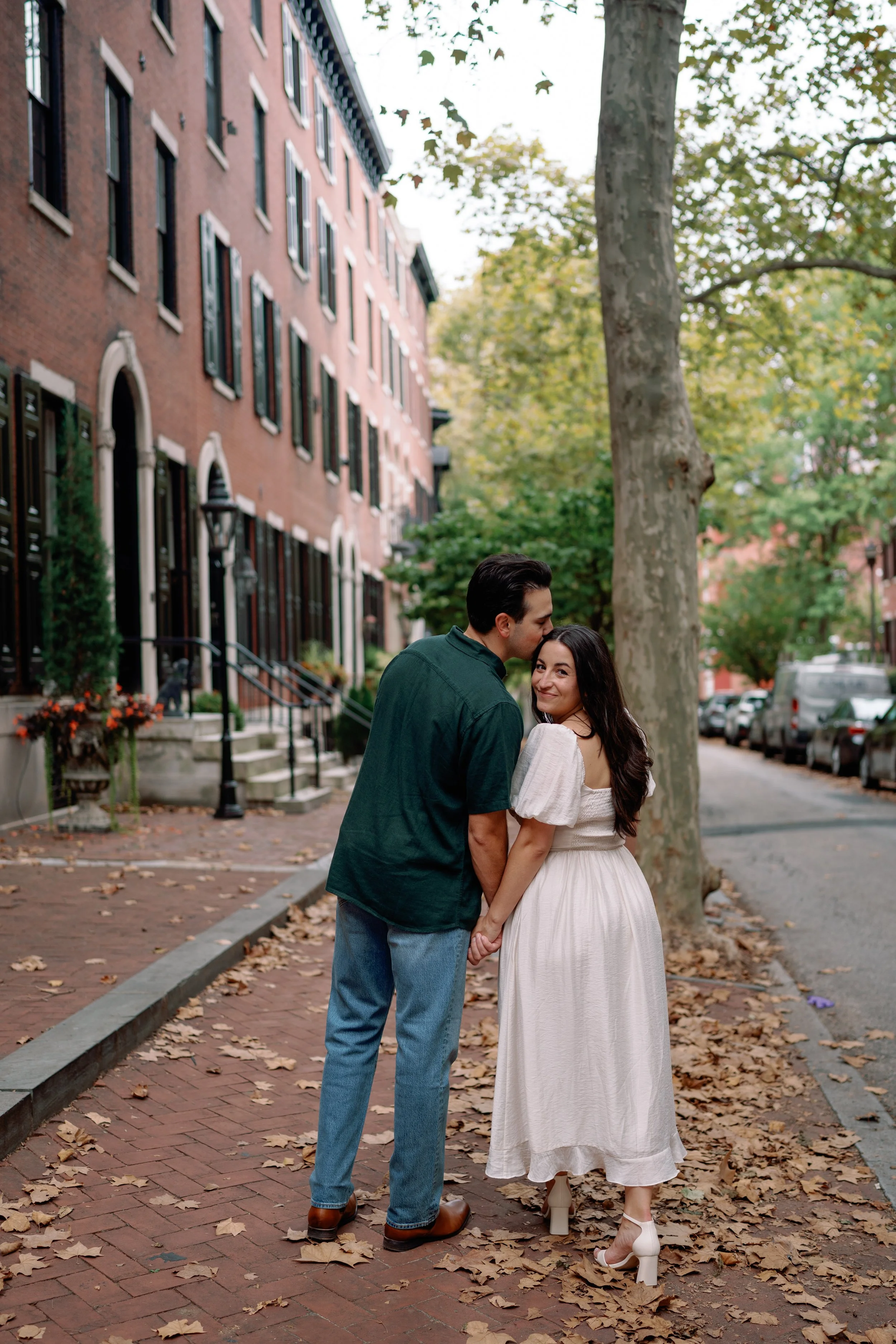 A couple holding hands and sharing a kiss on a fall sidewalk with fallen leaves, brick buildings, and trees in the background.