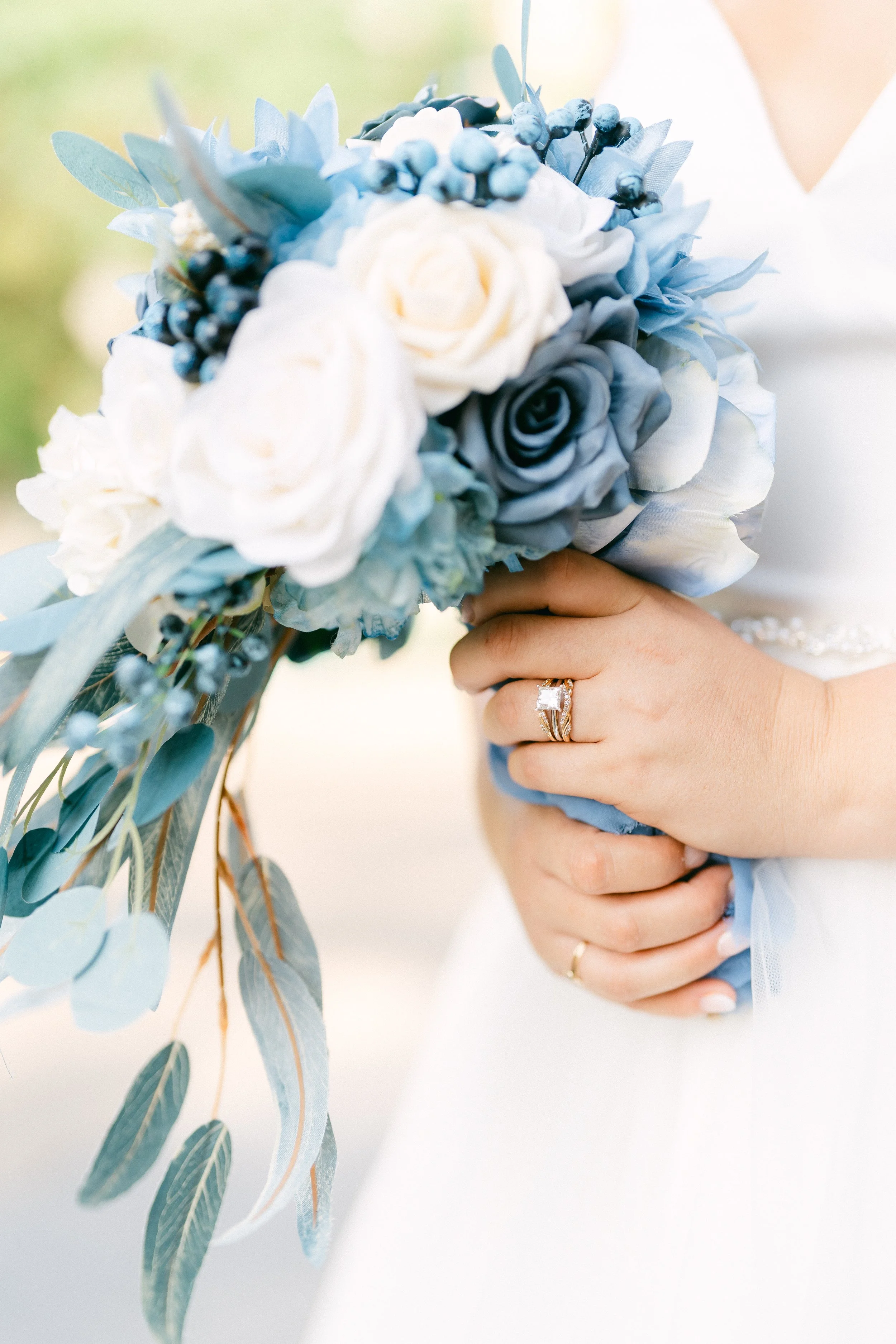 Close-up of a bride holding a blue and white floral bouquet, showing wedding rings on her fingers.