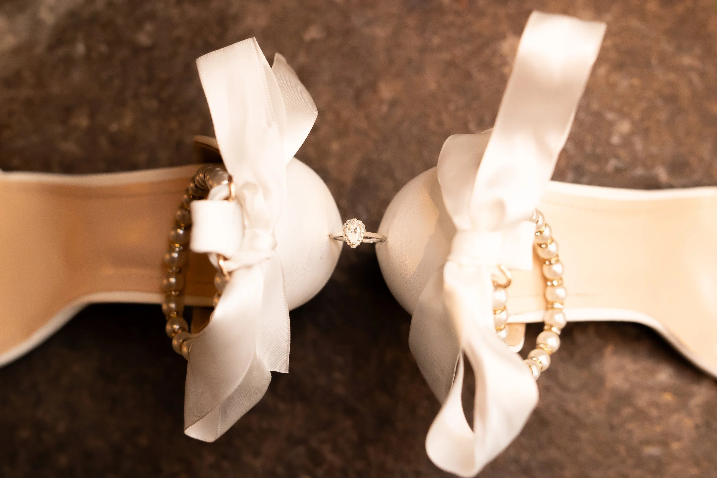 A diamond engagement ring between two white satin ribbon bows on white padded jewelry holders, with pearl bracelets draped over the bows.