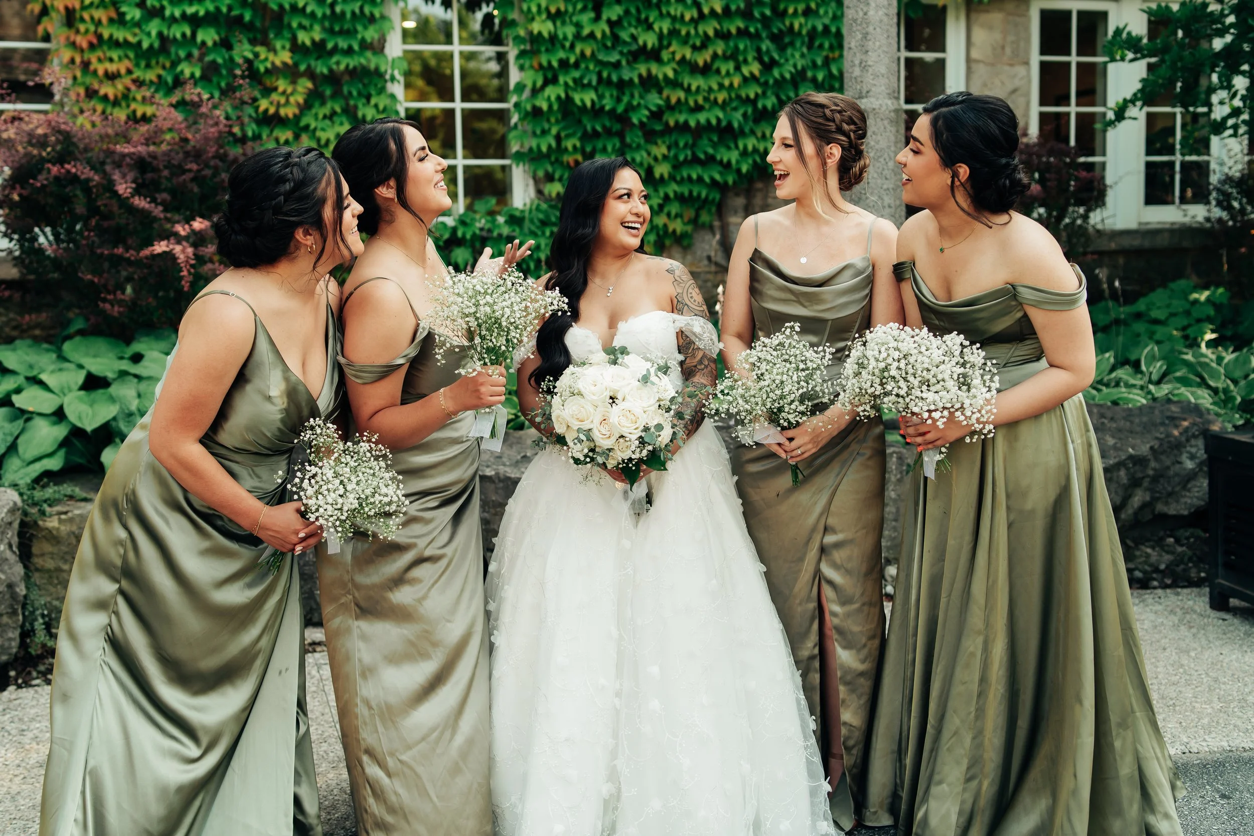 A bride and five bridesmaids standing outside in front of a green ivy-covered wall, smiling and talking, with each woman holding a bouquet of white baby's breath flowers.