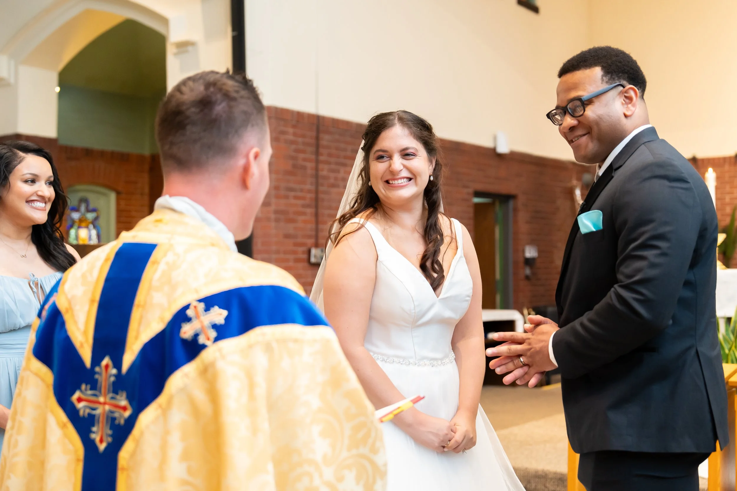 A bride smiling at a groom during their wedding ceremony inside a church with brick walls.