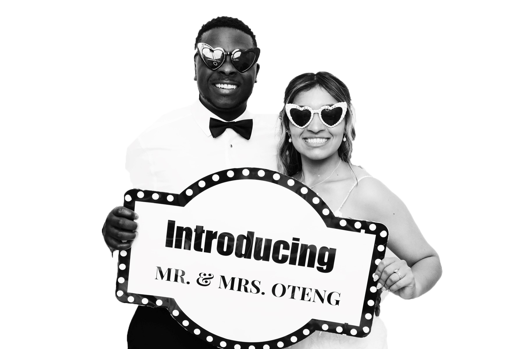 A smiling couple, dressed formally, holding a sign that says "Introducing Mr. & Mrs. Oteng," wearing sunglasses with heart-shaped frames, at a celebration event.