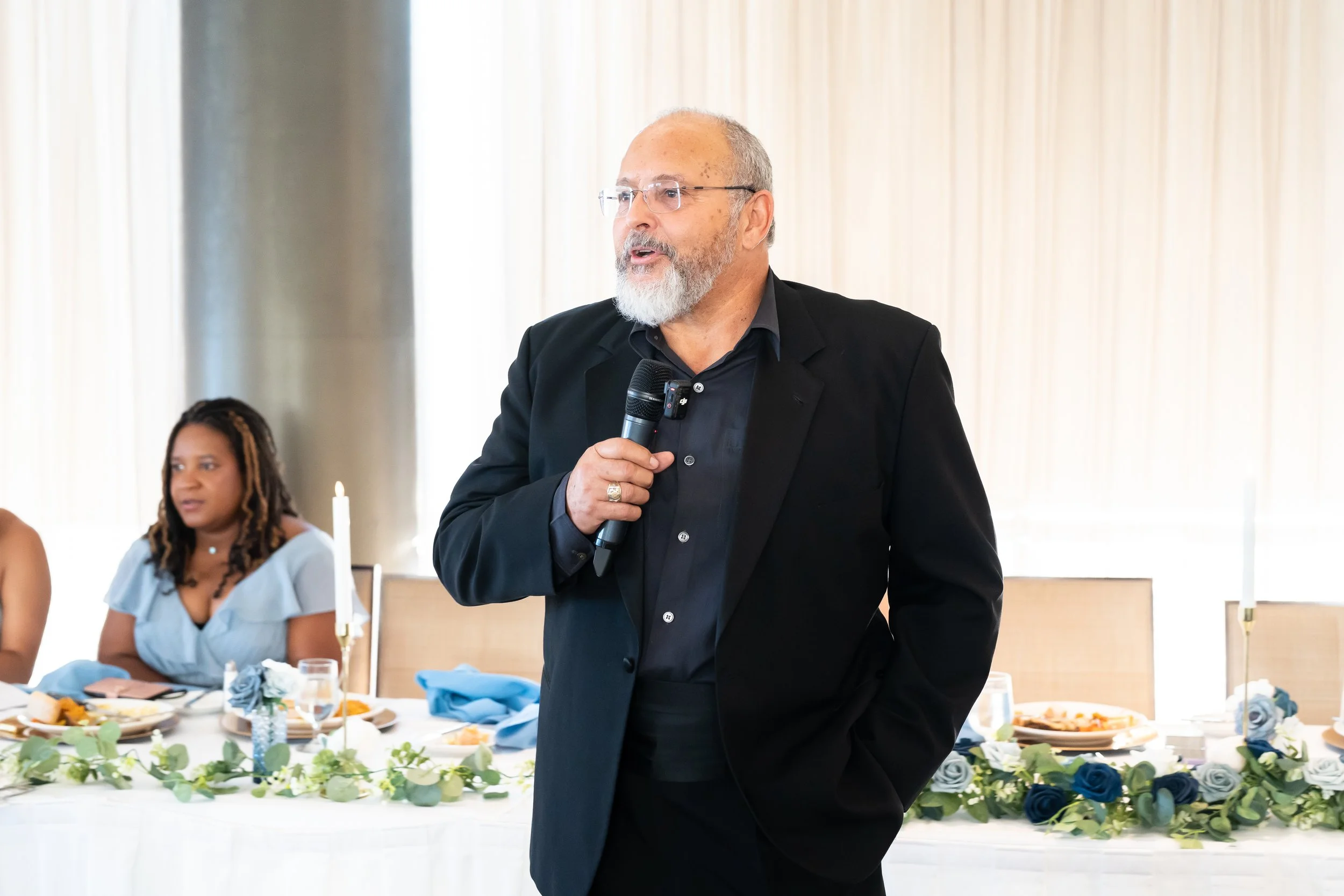 An older man with glasses and a beard, dressed in a black suit, holding a microphone and speaking at a formal event, with a woman seated at a decorated table in the background.