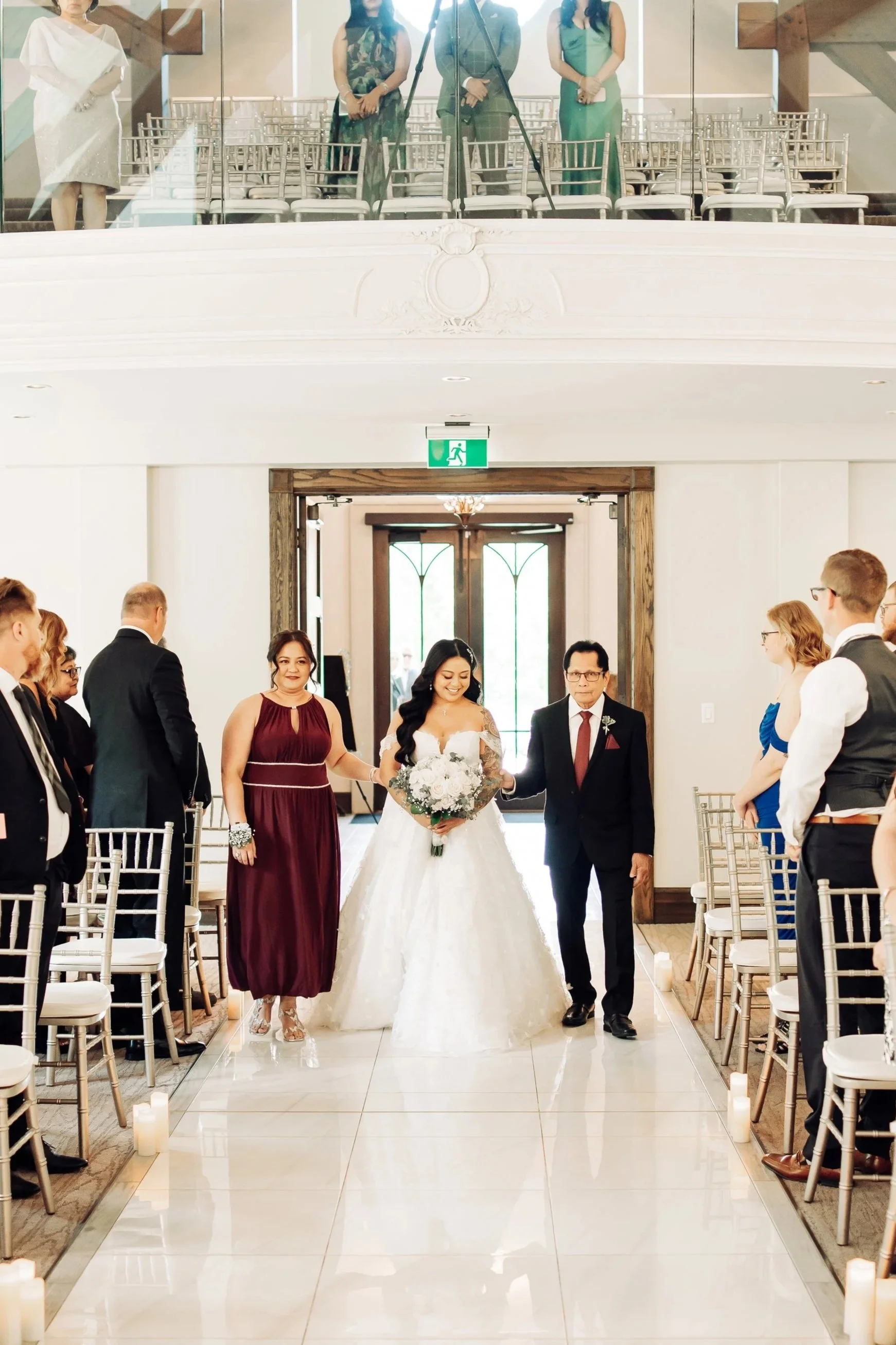 A bride in a white wedding gown walking down the aisle with her parents before the wedding ceremony, with guests seated on either side and candles along the aisle.