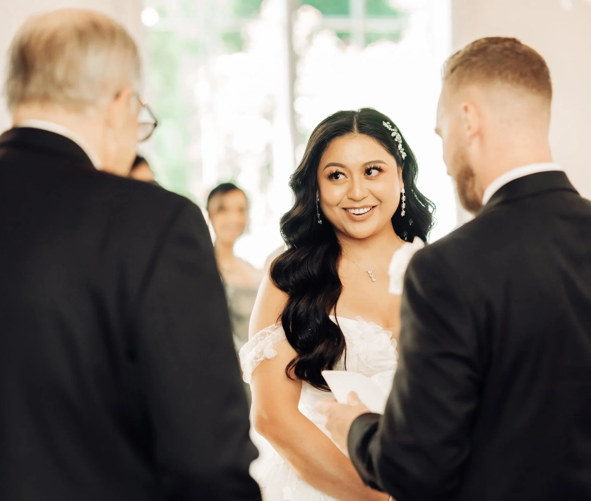 A bride and groom exchanging vows during a wedding ceremony, with an officiant present, indoors with natural light.
