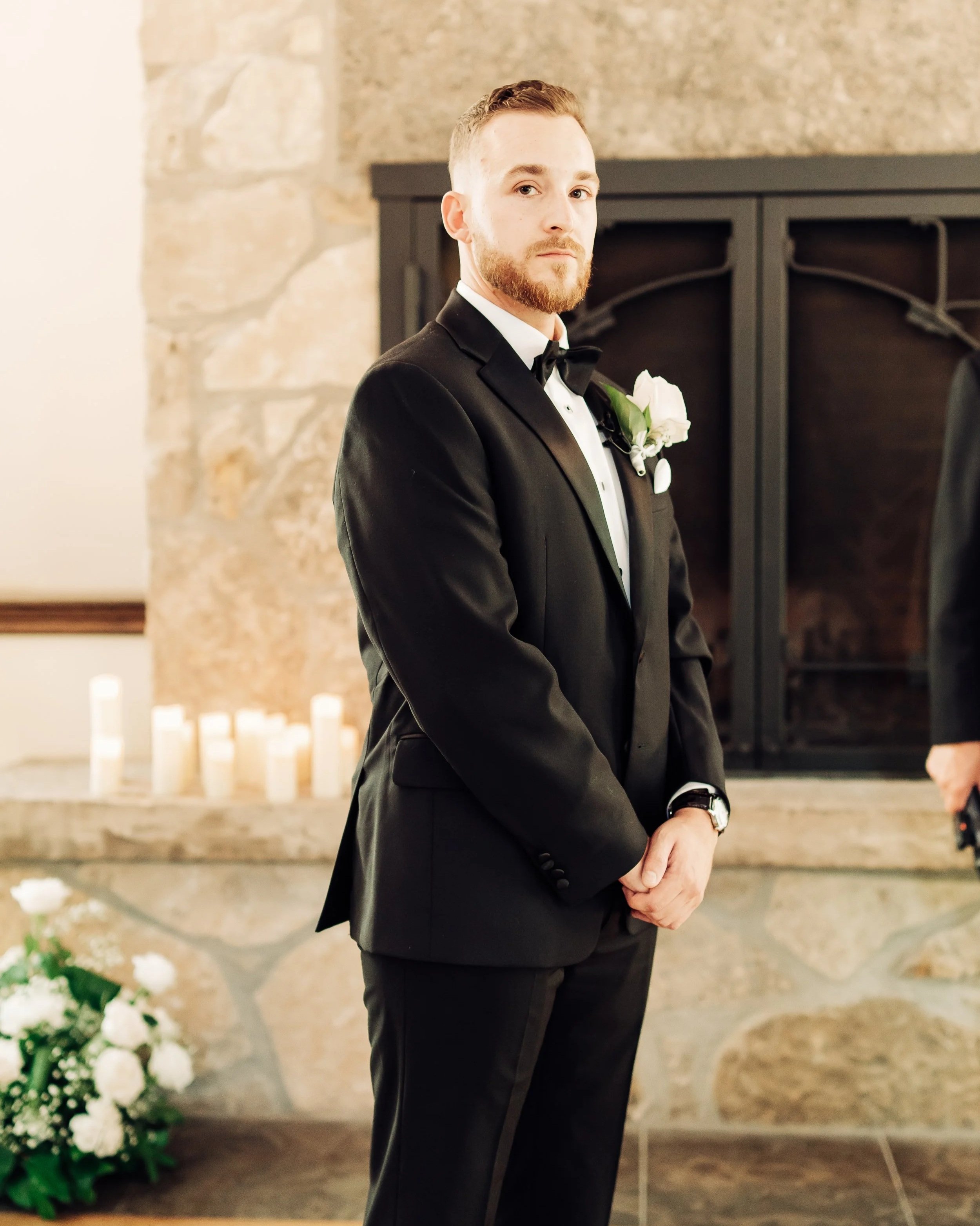 A man in a black tuxedo with a white shirt and black bow tie standing indoors near a stone fireplace, with candles and white flowers in the background.